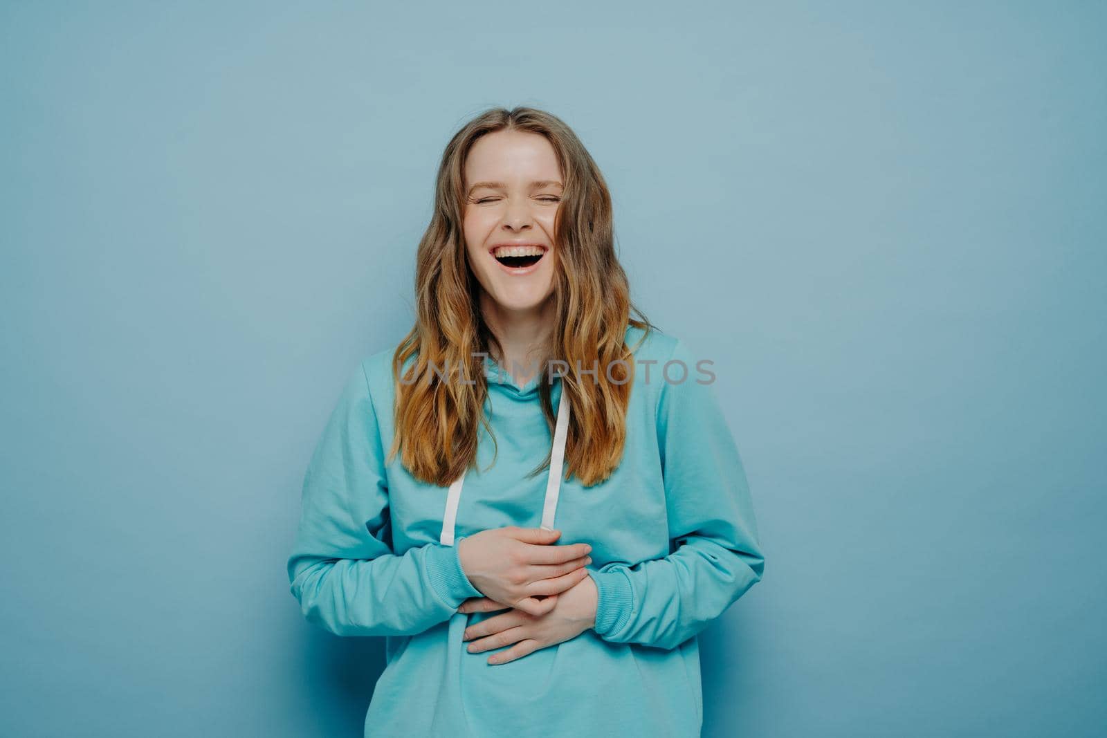 Overjoyed young woman dressed in hoodie holding belly and cant stop laughing after hearing hilarious joke, keeping eyes closed and having fun while standing isolated over blue background, studio shot
