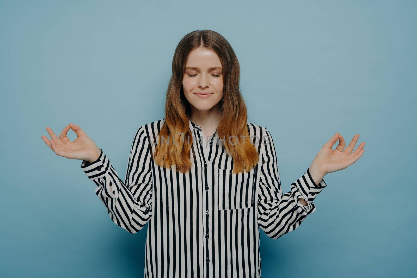 Young peaceful female student in casual clothes meditating with closed eyes, holding hands in yoga zen gesture, feeling calm and positive, teenage girl relieving from stress. Studio shot. Life balance