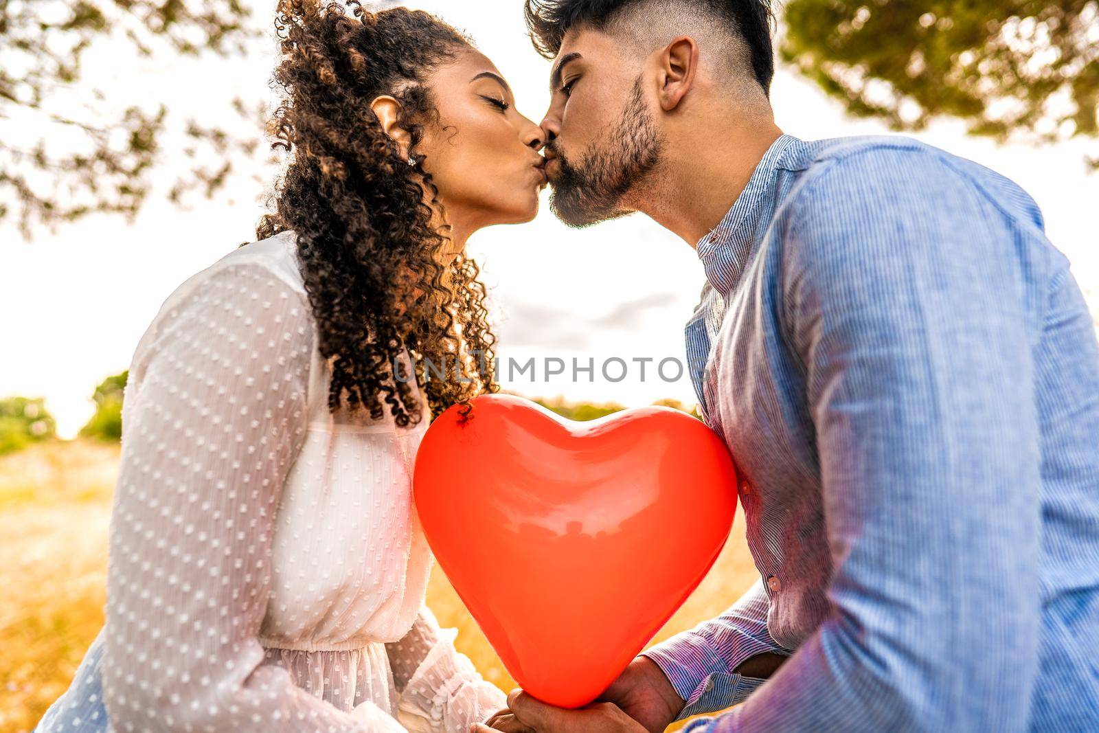 Profile photograph of multiracial couple in love kissing at sunset in nature with sun backlit effect on red heart shaped balloon among them. Romantic scene at dusk of two heterosexual young people