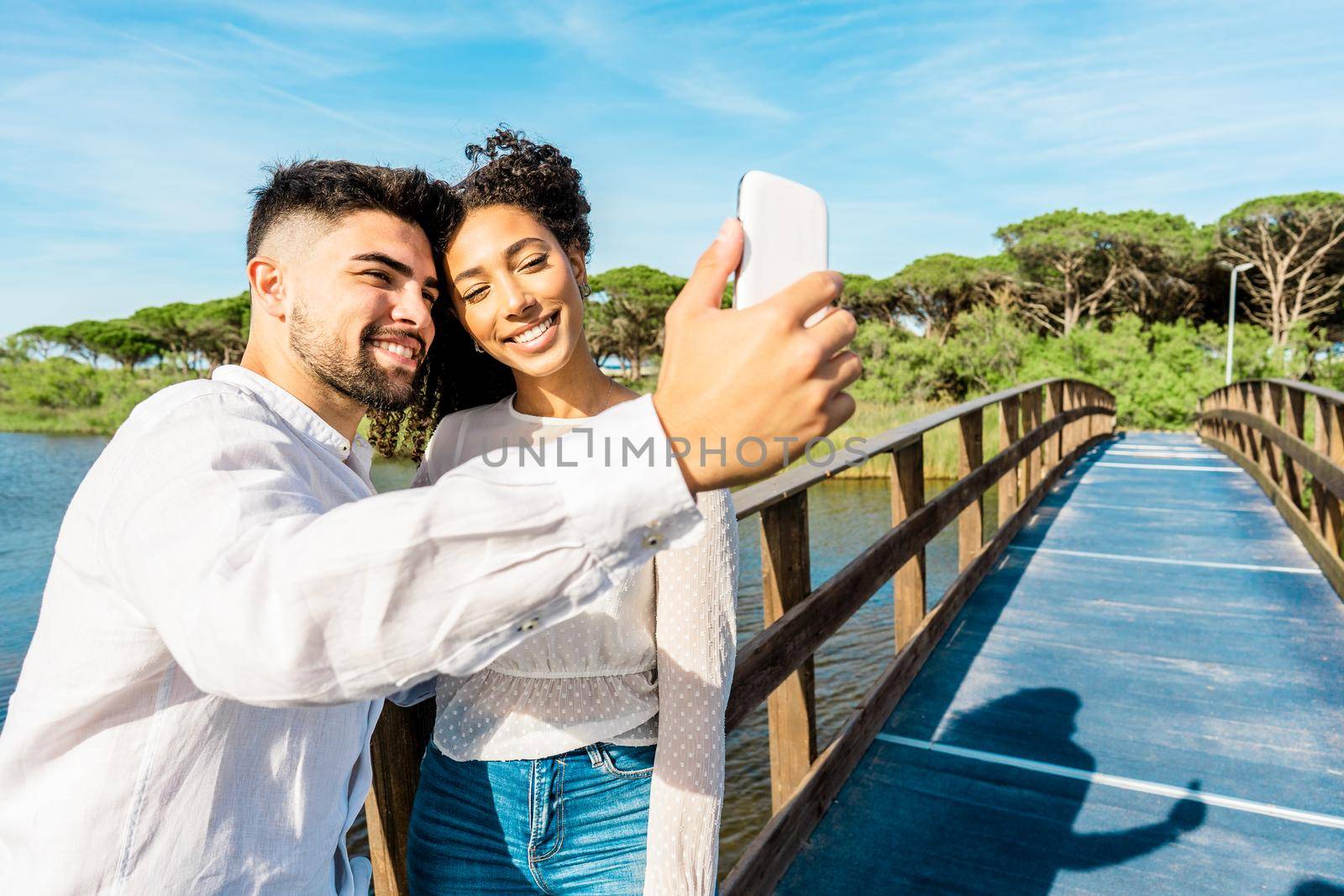 Happy multiracial couple in love staying on a wooden bridge taking a selfie in nature vacations. Handsome guy photographing himself with Hispanic girlfriend using smartphone. New technology habits