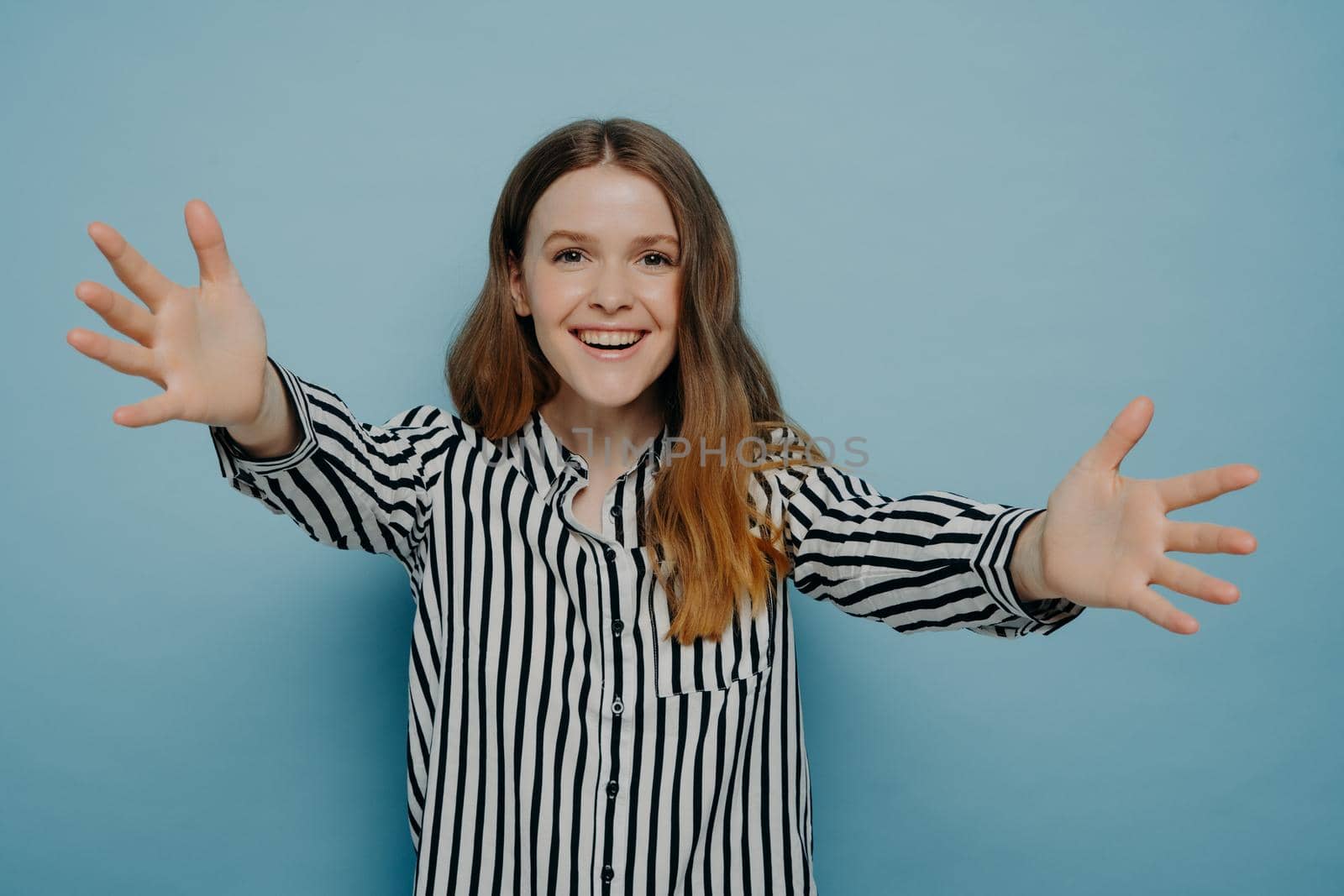 Portrait of loving young woman offering hug, smiling and pulling hands towards camera while standing isolated on blue background, cheerful teenage girl going to cuddle best friend