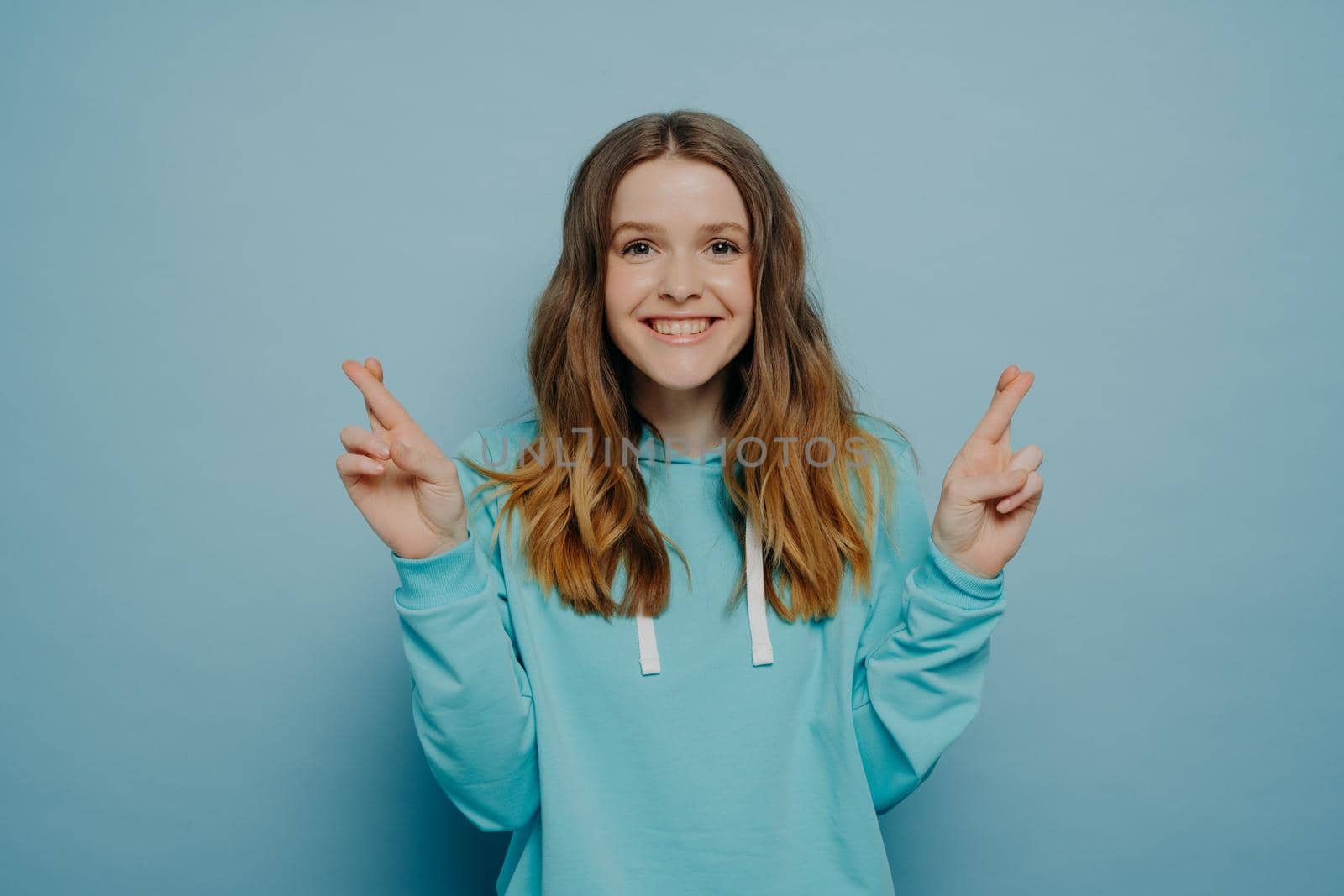 Excited smiling young girl with wavy ombre hair keeping fingers crossed looking at camera wearing casual sweater posing isolated on light blue studio background. Body language concept