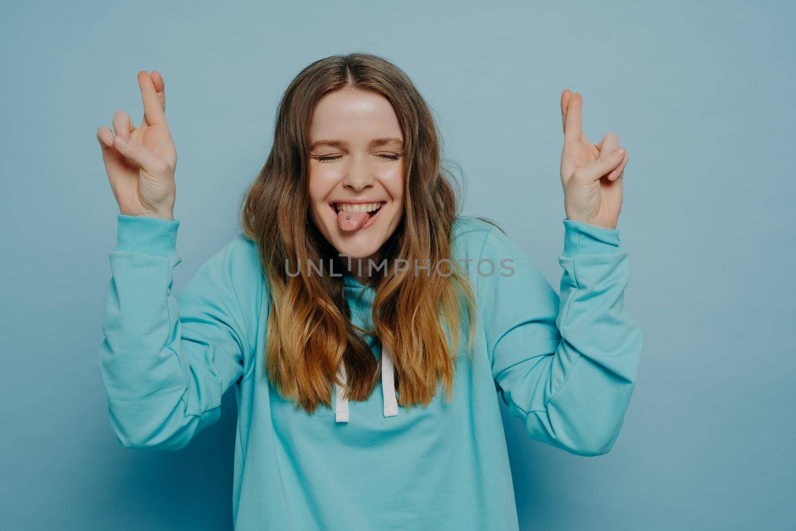 Happy crazy teenage girl with wavy ombre hair keeping fingers crossed while closing eyes and sticking tongue out standing against blue wall in studio, having fun and feeling carefree