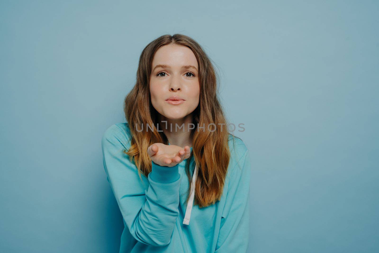 Pretty young girl sending air kiss posing over blue background by vkstock
