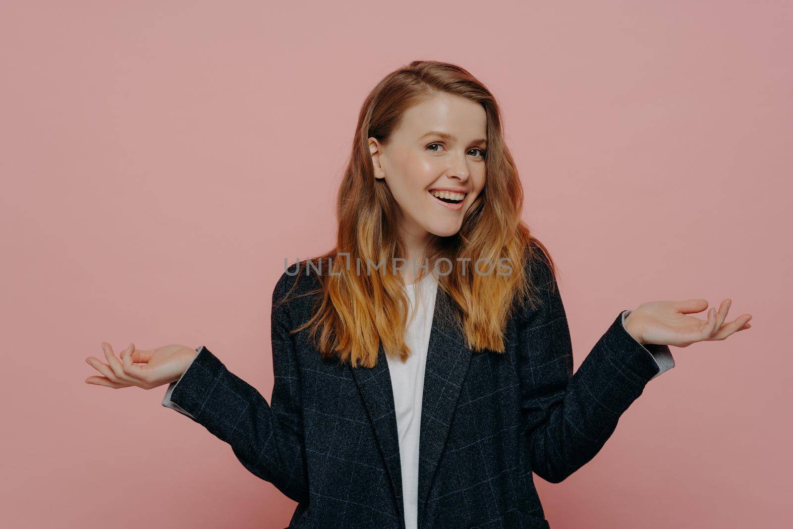 Happy attractive young woman with thick medium length ginger hair wearing dark formal jacket and white top raising hands to sides, indoor isolated on pink background. Body language concept