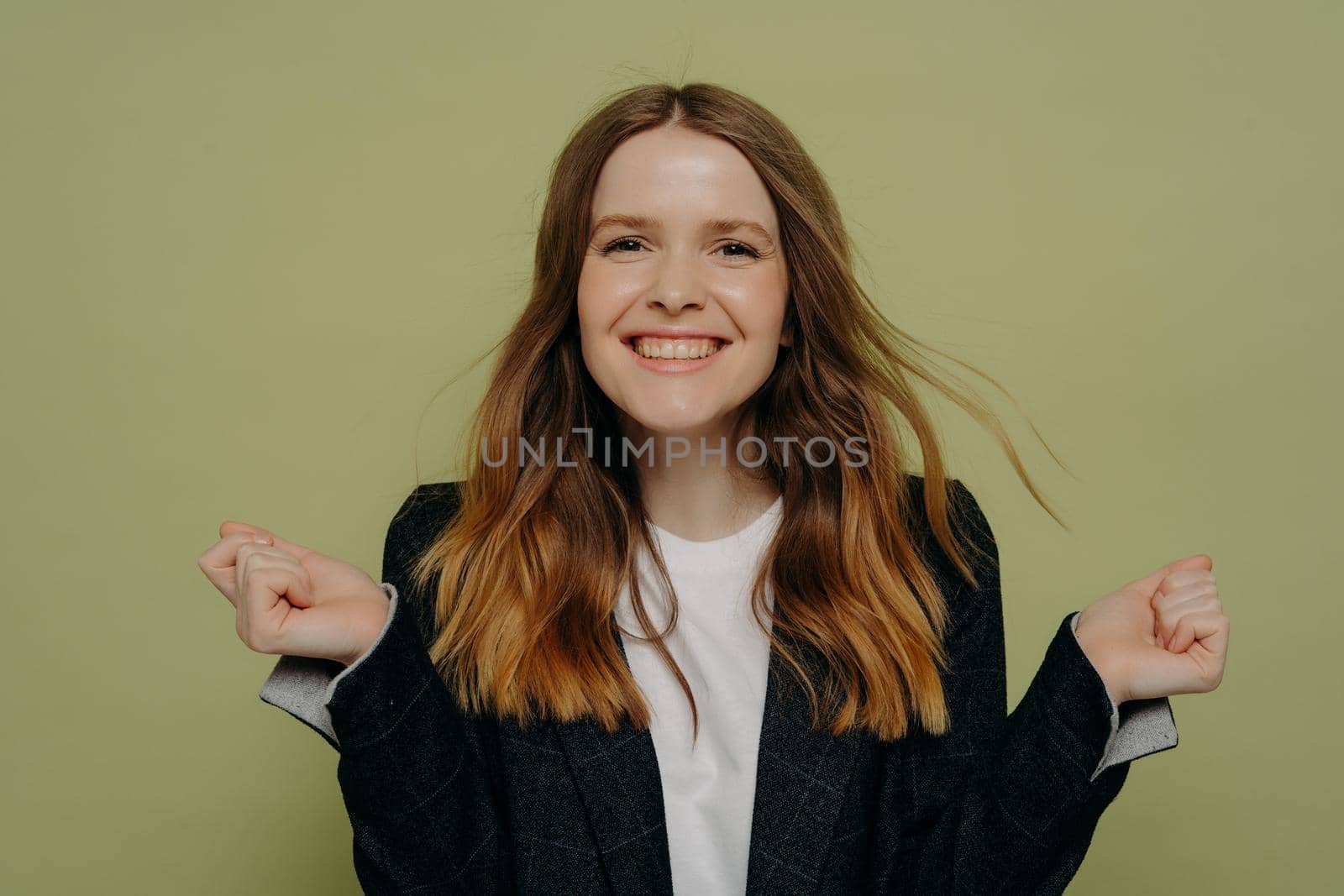 I did it. Smiling young woman holding hands up demonstrating excitement while looking at camera wearing dark jacket and white top, posing against light studio background. Human emotions concept