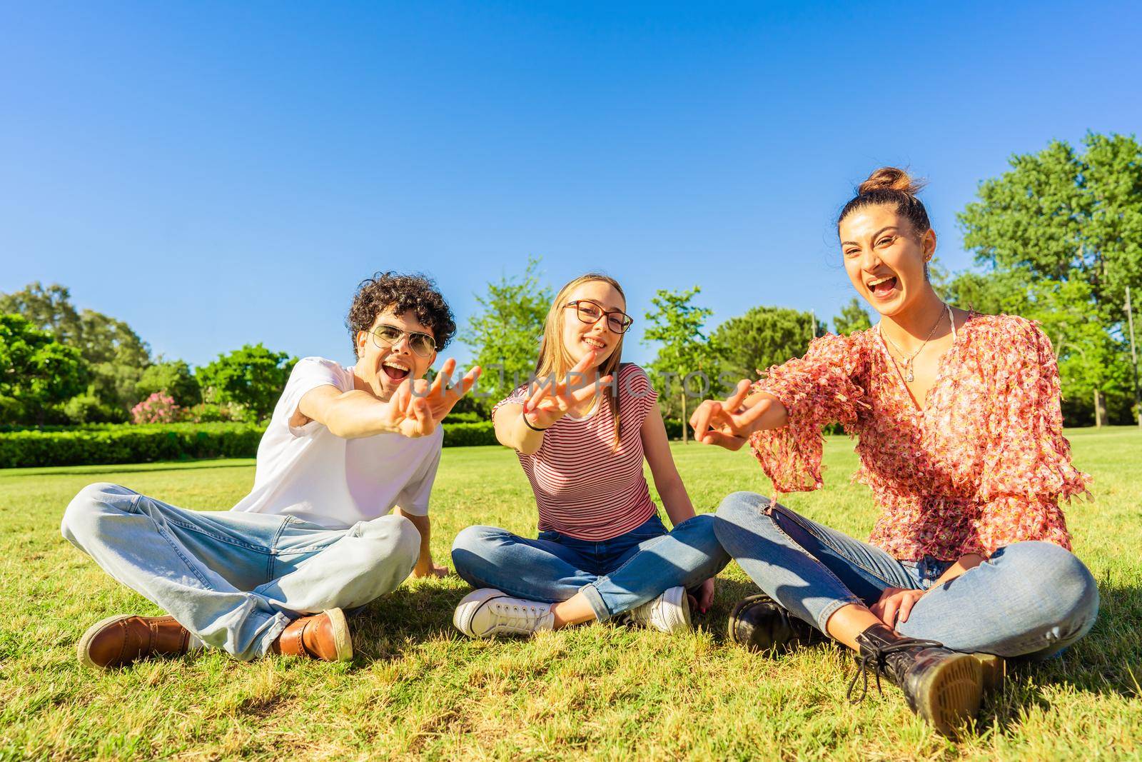 Three young student best friends sitting on grass in city park showing victory sign with two fingers looking at camera. Concept of unity and solidarity in youth age. Happy gen z smiling in nature