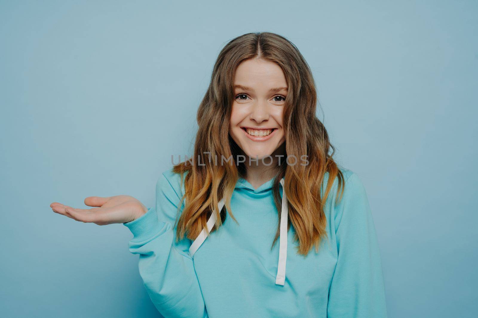 Smiling teenage girl with tipping hand shrugging shoulders standing against light blue background, cute female with wavy ombre medium length hair raising eyebrows. Human emotions concept