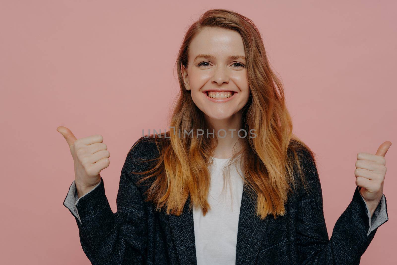 Confident smiling young woman with wavy ginger hair showing thumbs up with both hands, wearing dark formal jacket and white top posing on light pink background. Body language concept