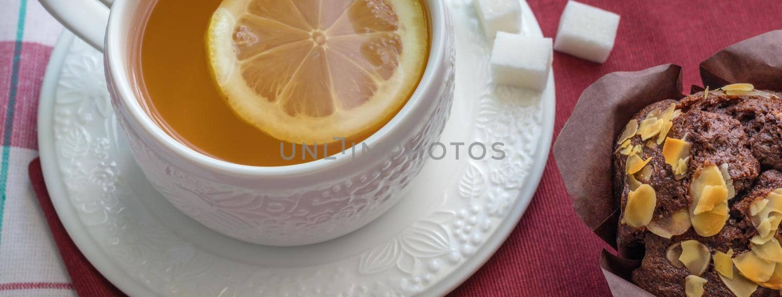 Tea with lemon in a white cup on a saucer with cubes of raffinated sugar and chocolate muffin with pistachios on a red napkin. High angle view, panoramic size