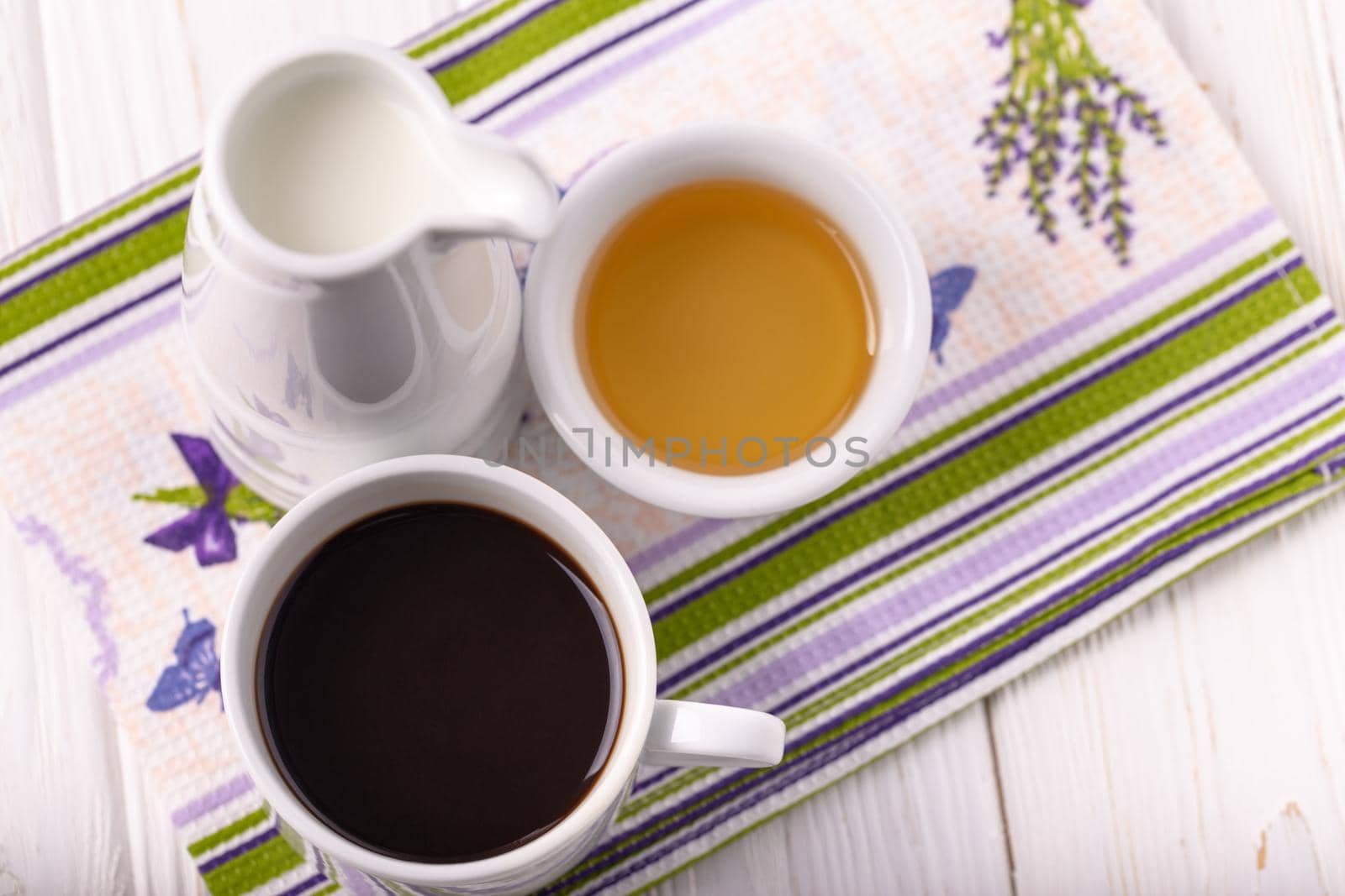 A cup of coffee, milk in a jar and honey in a bowl on a colorful napkin. White wooden background, rustic style, top view