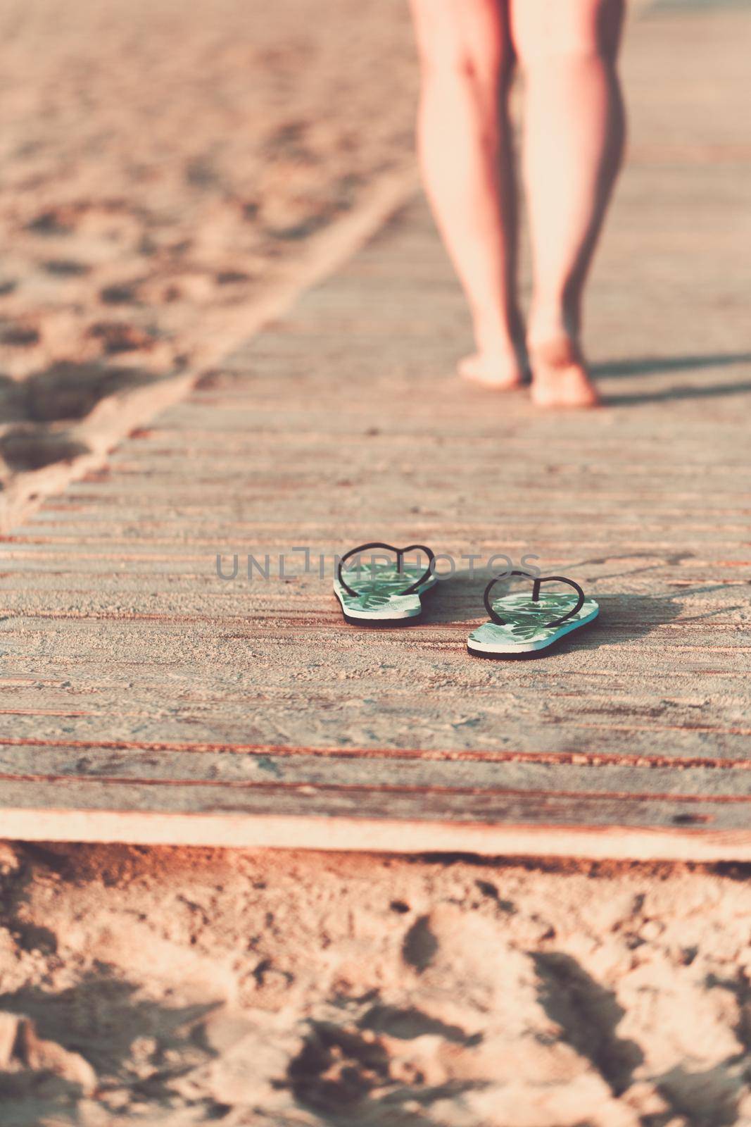 Bare feet of a girl walking on a wooden floor on the seashore at sunny summer morning. Green flip flops in the foreground.