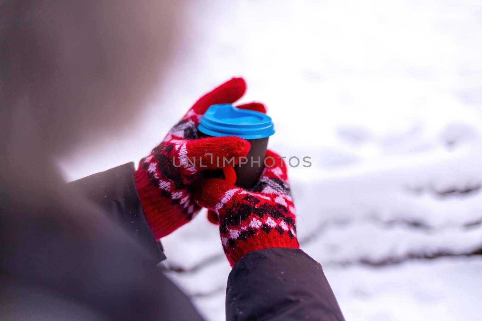 Women's hands in a red mittens hold a paper cup of coffee. Back side view on white snowy background.