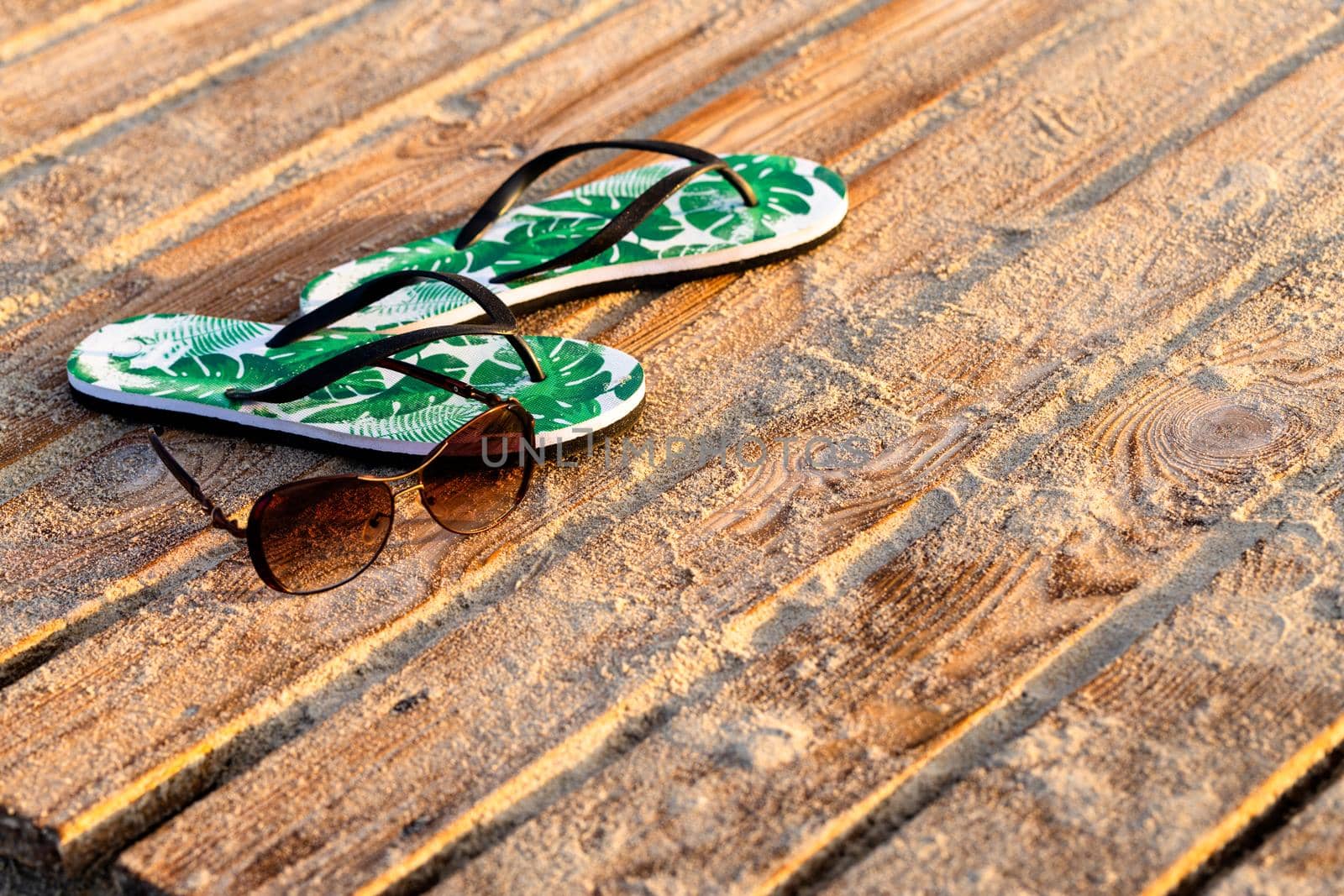 Green flip flops and sunglasses on the wooden boards covered with the beach sand. Close-up, early morning golden sunrise
