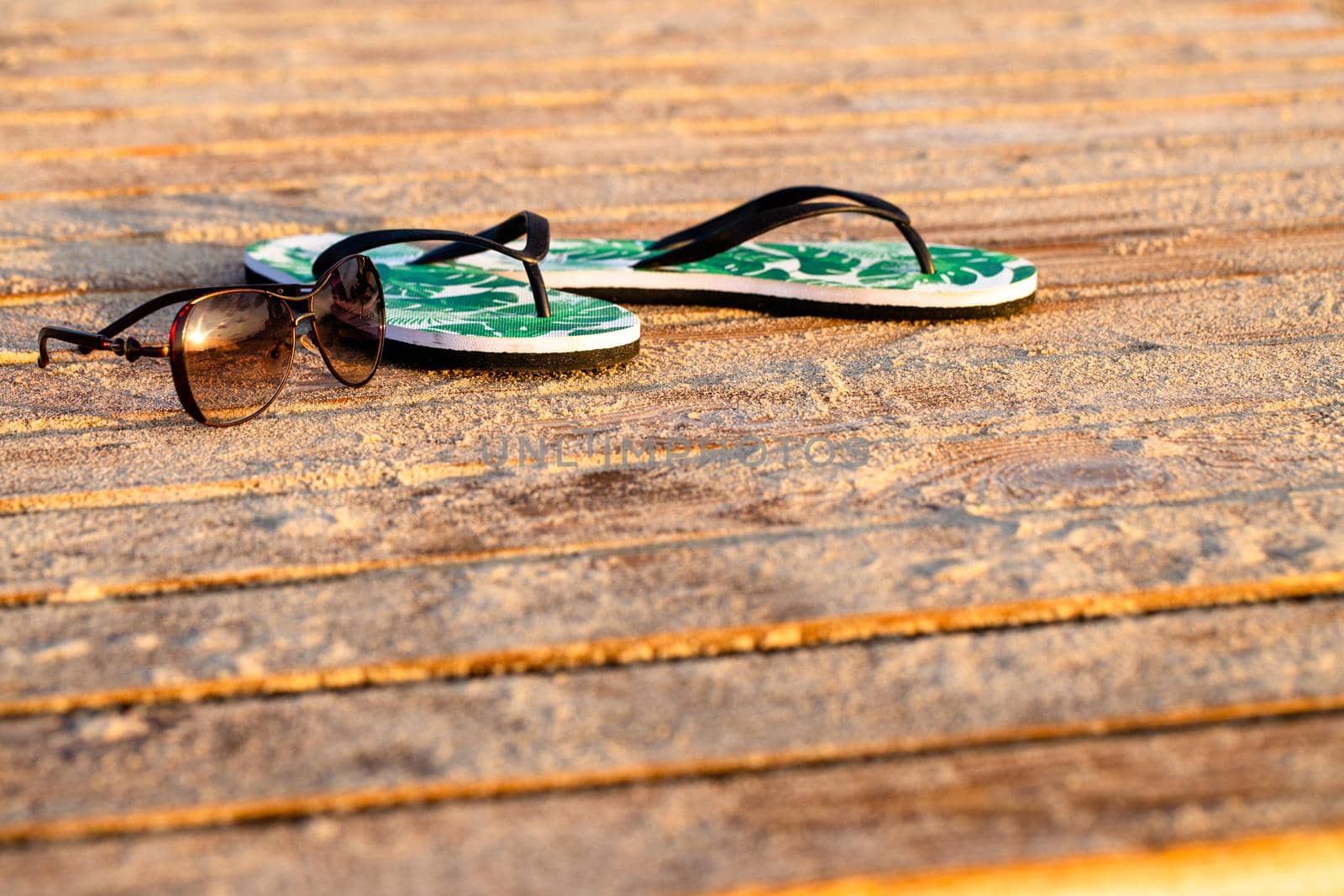 Green flip flops and sunglasses on the wooden boards covered with the beach sand. Close-up, early morning golden sunrise
