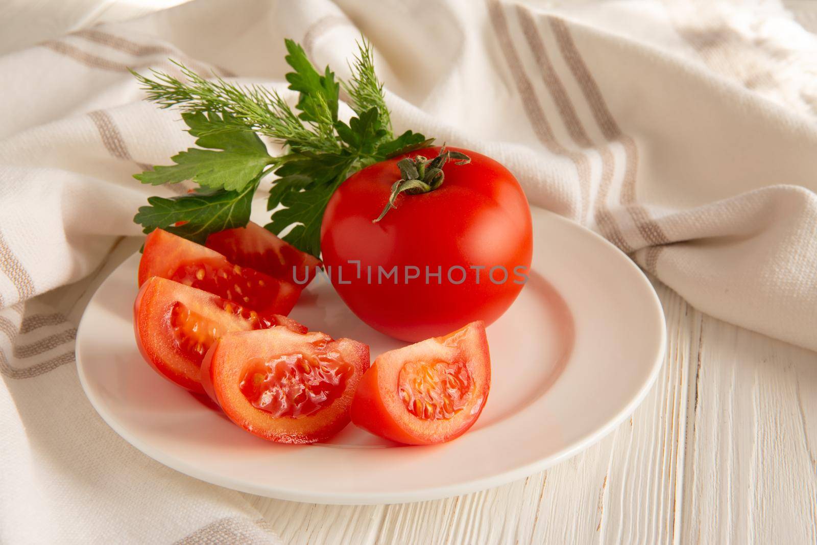 Ripe red tomatoes with a bunch of green parsley on a ceramic plate in a linen towel under soft sunlight