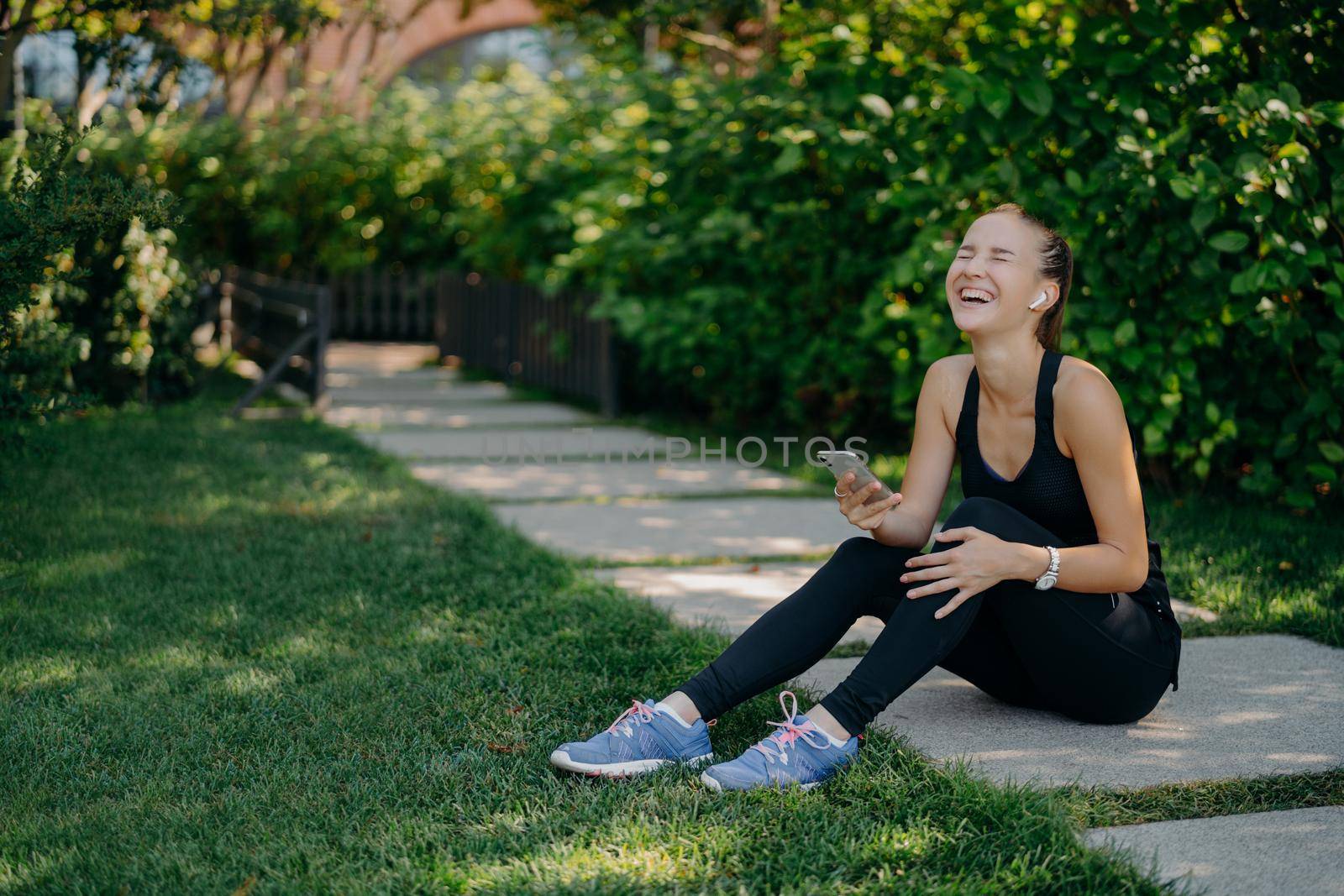 Overjoyed sportswoman sits on path laughs out uses smartphone for listening music from playlist share content and chatting dressed in active wear enjoys spare time. Active lifestyle concept.