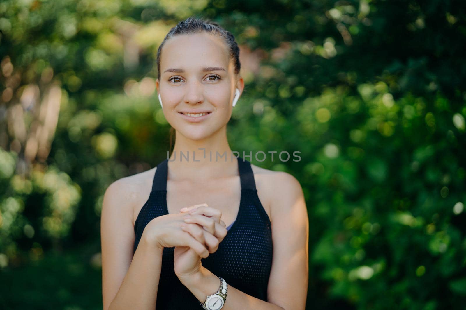 Outdoor shot of pretty dark haired woman keeps hands together being in good physical shape as goes in for sport regularly wears sportsclothes listens music while having workout at green park by vkstock