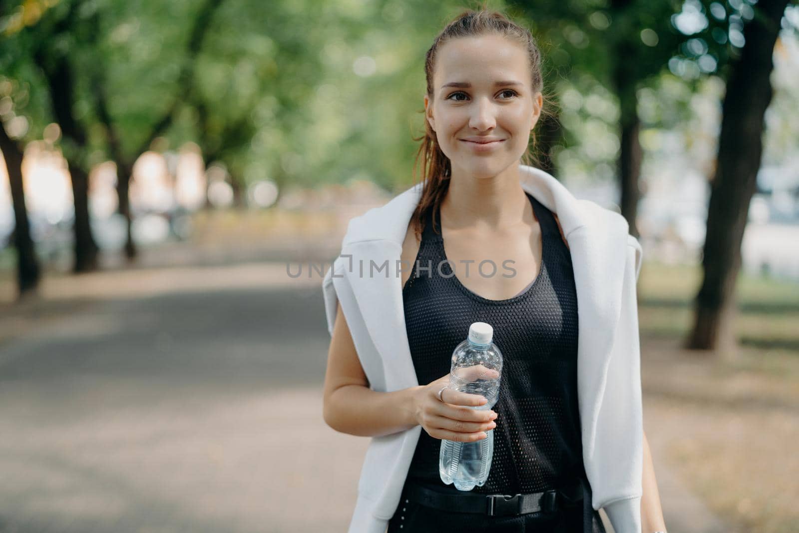 Cheerful young female runner holds water bottle poses outdoor in active wear takes break after workout walks in park gets refreshment after jogging feels thirsty keeps fit with regular sport