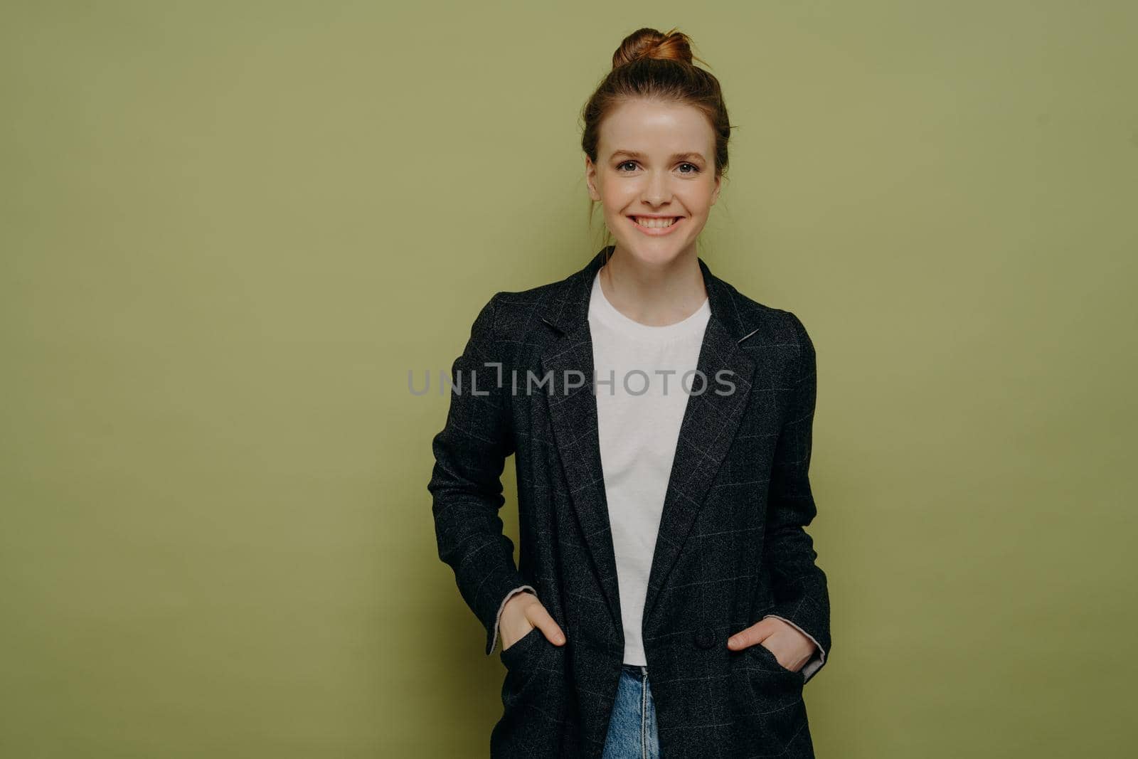 Smiling confident teenage girl with hair in bun wearing stylish plaid blazer and jeans keeping hands in pockets and looking at camera with happy expression while standing against green wall