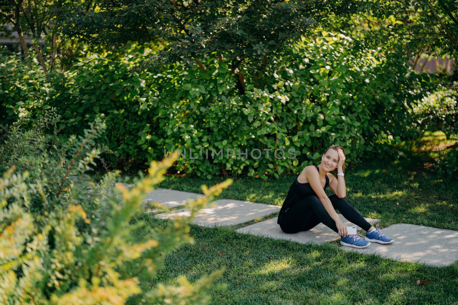 Horizontal full length shot of pleased brunette sporty woman listens motivation playlist before start running dressed in active wear poses on path uses application online. Sport and wellness concept by vkstock