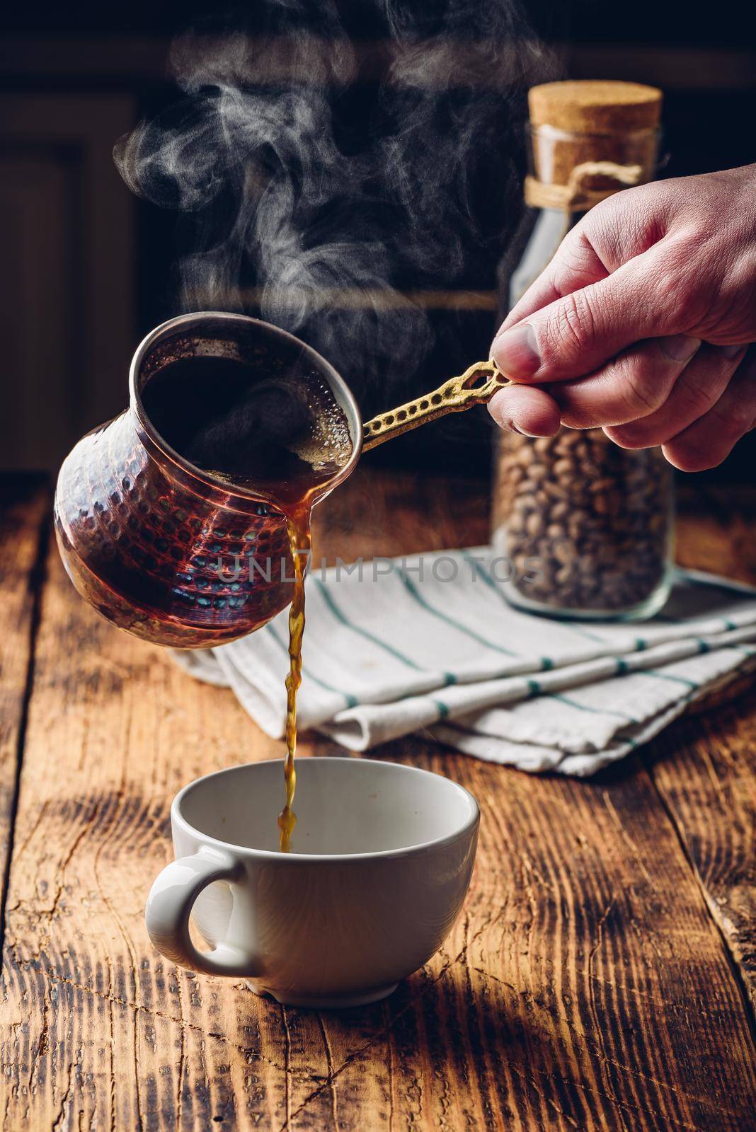 Pouring turkish coffee into white cup. Jar of roasted coffee beans on background