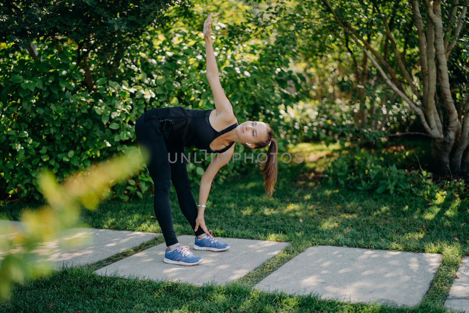 Active fitness model exercises outdoors leans to feet raises arm prepares muscles before cardio training challenges herself dressed in active wear has pony tail poses in green park during sunny day by vkstock