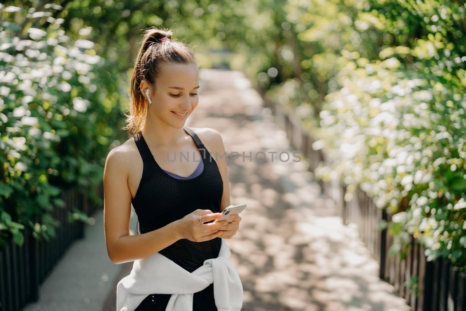 Sport as style of life. Horizontal shot of healthy slim sporty woman focused at smartphone checks information and chooses song from playlist dressed in sportwear poses outdoor breathes fresh air by vkstock