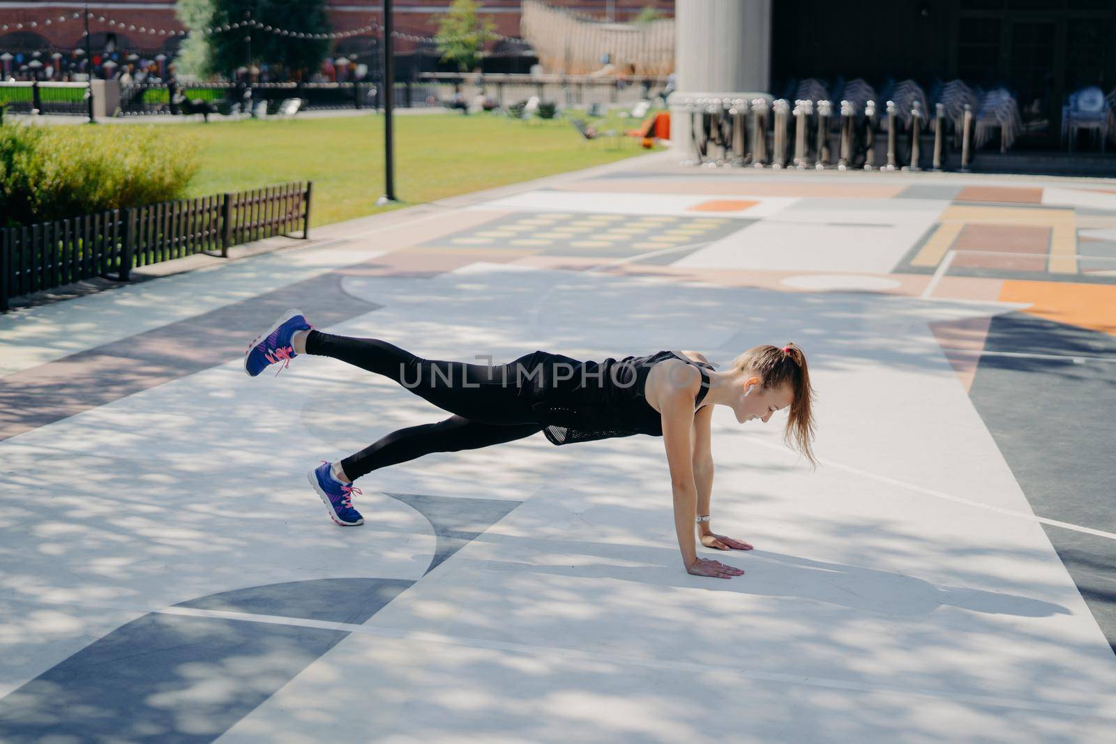Sporty slim young woman does planking exercise raises leg demonstrates her determination wears sportsclothes and sneakers trains outdoors listens music in earphones. Healthy lifestyle concept by vkstock