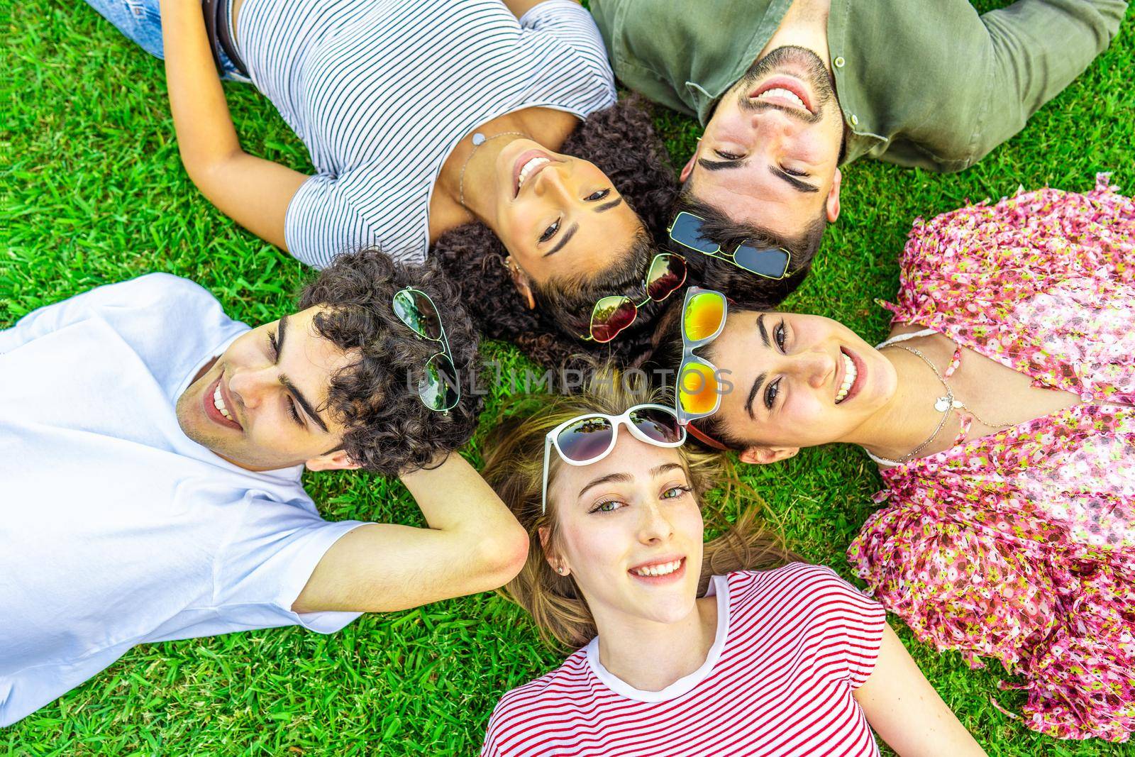 Shot from above of five happy multiracial friends lying head to head with colored sunglasses looking at camera from bottom. Mixed race group of smiling cute young people having fun together in nature by robbyfontanesi