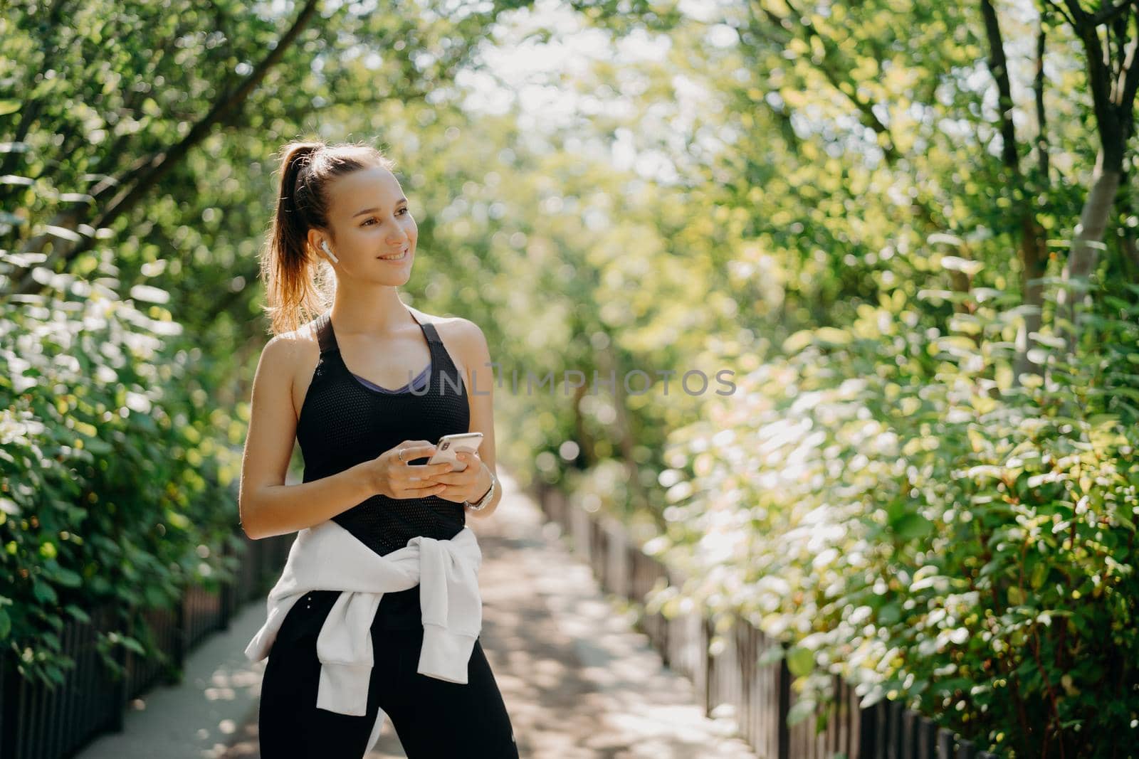 Horizontal shot of active sporty woman uses smartphone for checking results after jogging dressed in active wear enjoys warm sunny day listens music via earphones. Healthy lifestyle concept. by vkstock