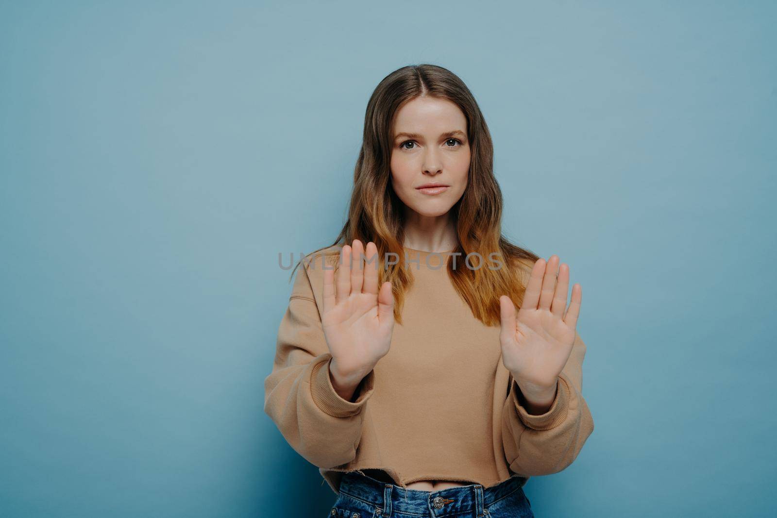 Serious attractive young woman with brown hair in casual wear holding hands in front of her demonstrating stop gesture while posing on blue light blue studio background