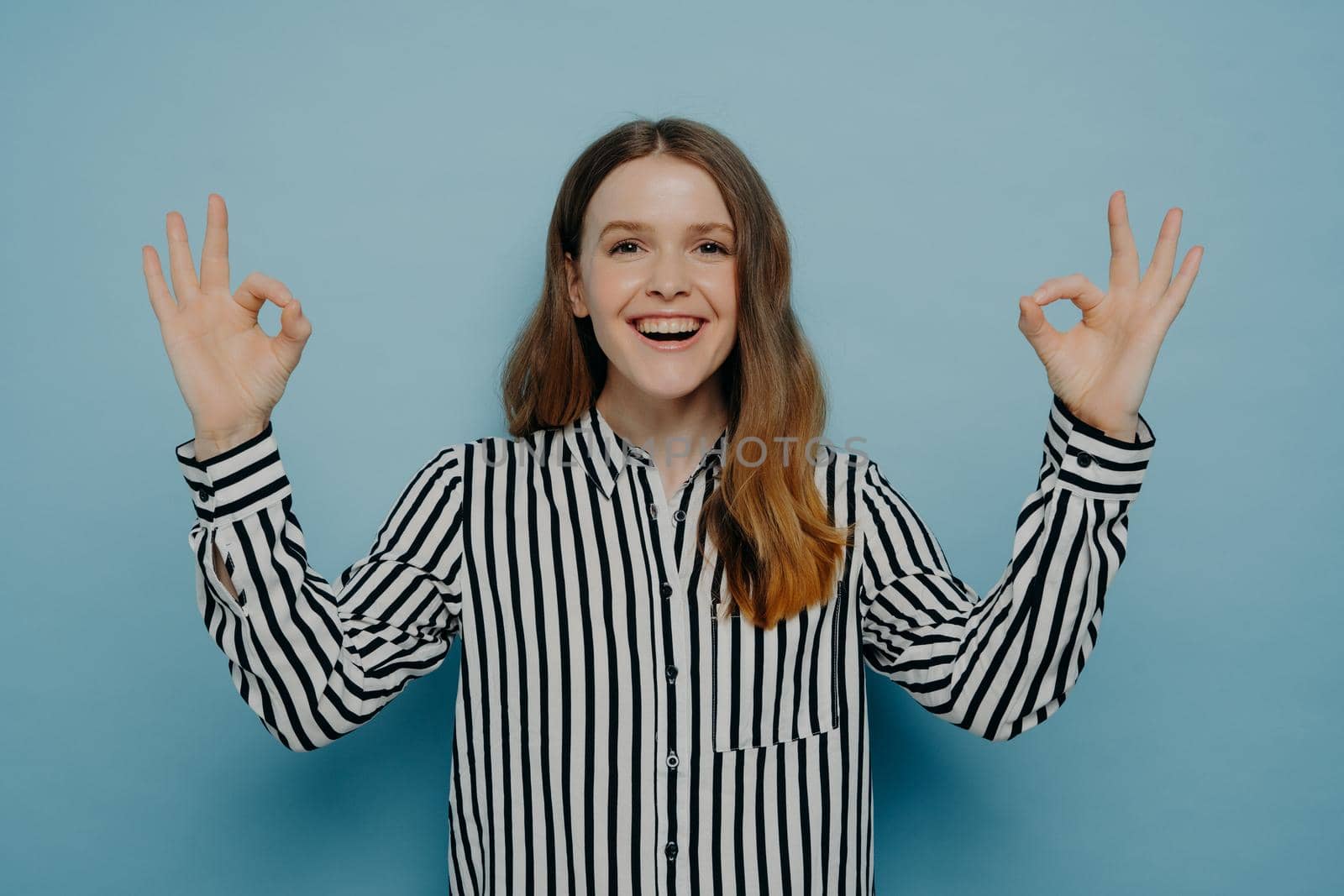 Laughing young female with wavy brown hair showing okay sign with both hands wearing striped black and white blouse while standing against blue wall in studio. Body language concept
