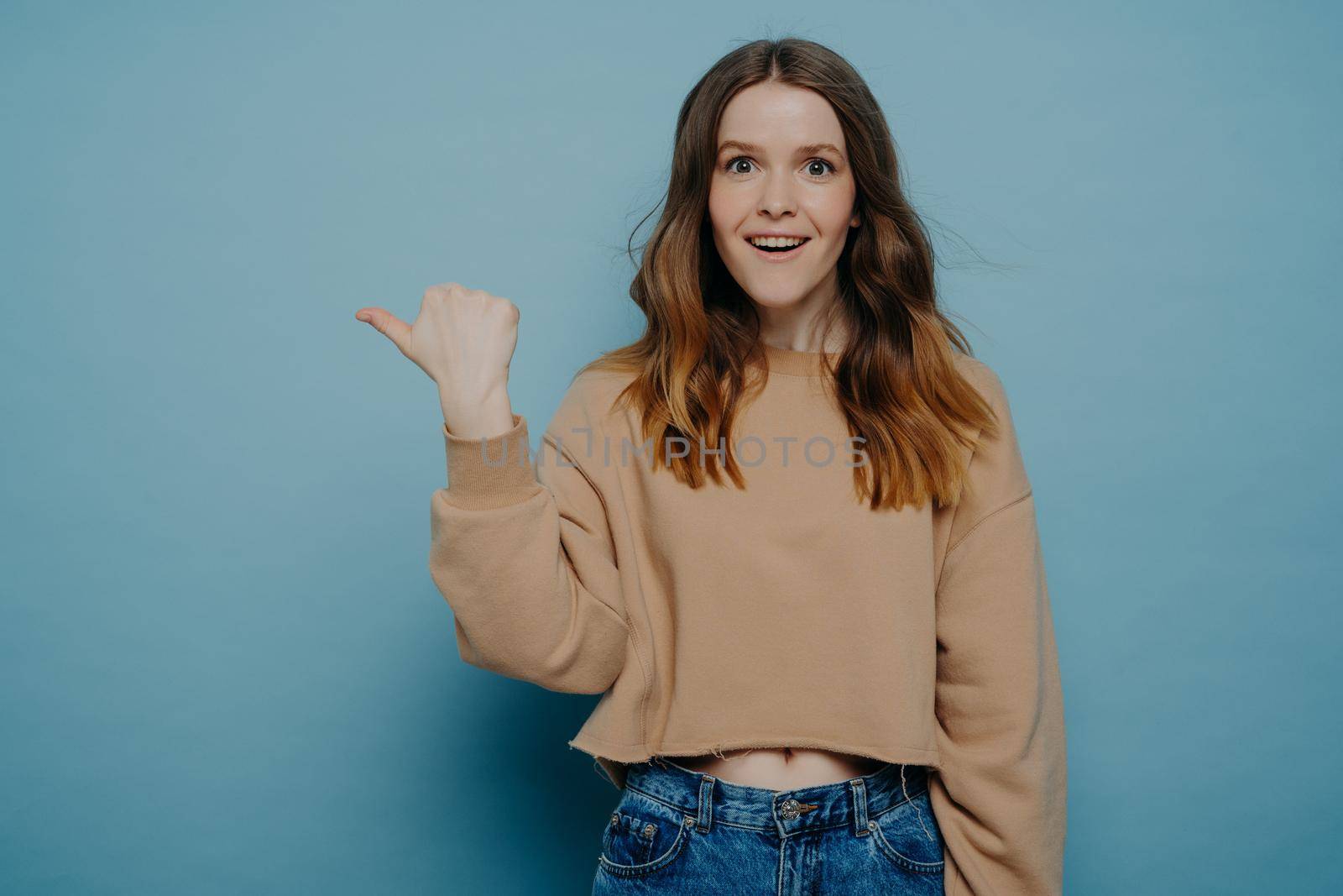 Amused pretty young female with ombre hair in loose casual light brown sweater and jeans pointing her finger at something attracting attention, standing against blue wall. Advertisement and promotion