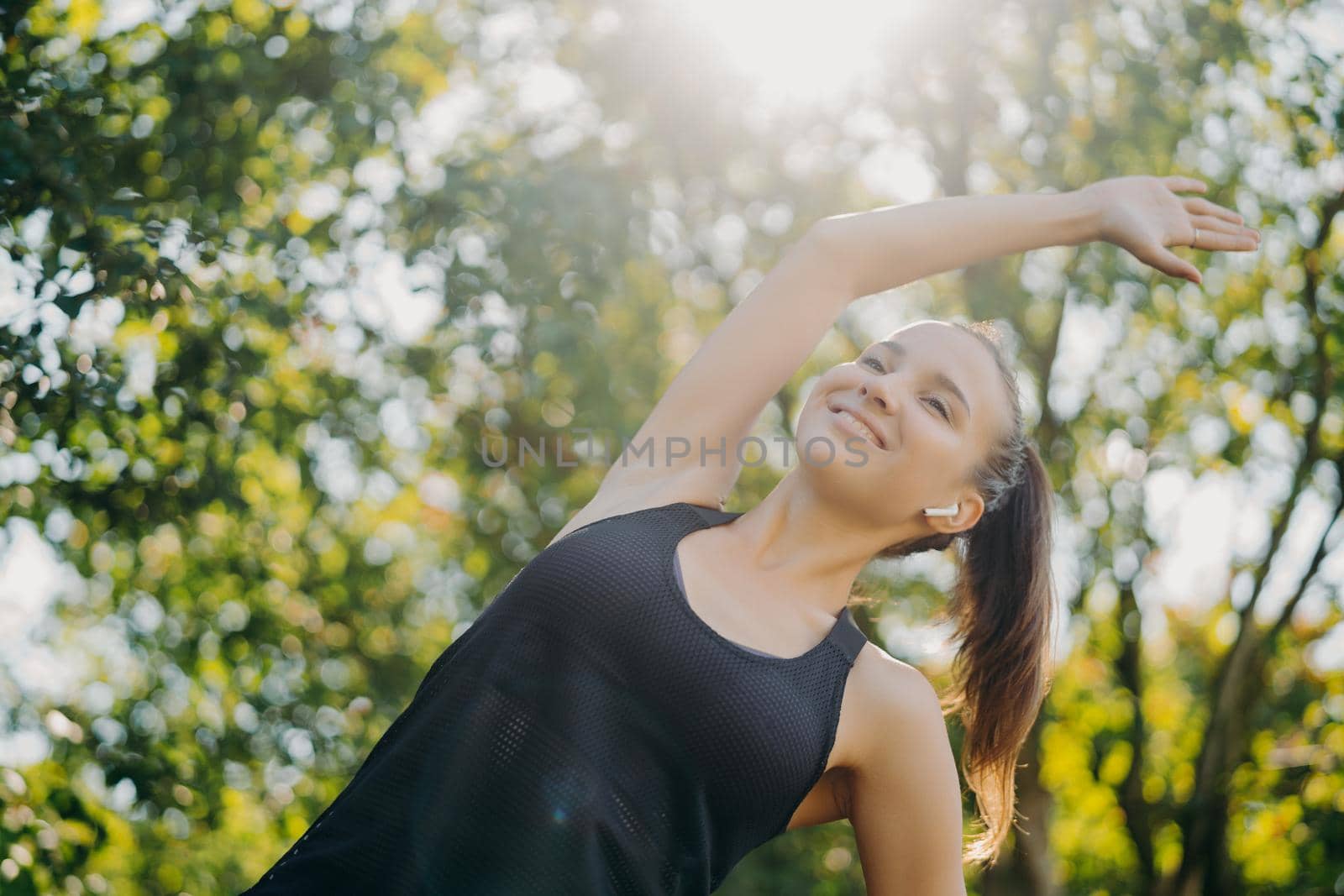 Cheerful sporty woman with pony tail leans aside exercises outdoor against trees enjoys sunny day listens music via wireless earphones has morning training seession performs warm up cardio workout