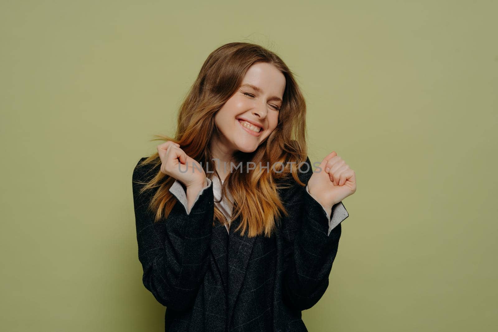 Celebrating success. Pleased young female with wavy brown hair holding hands up expressing happiness with closed eyes wearing dark formal jacket, posing against light studio background