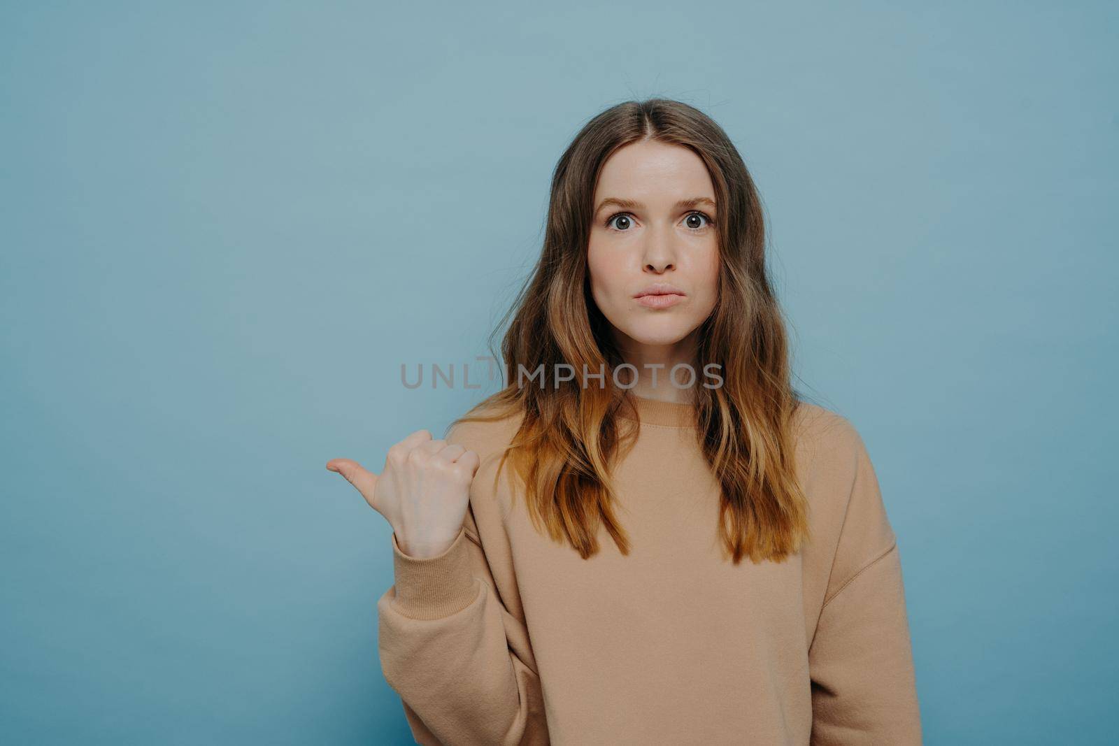 Surprised teenage girl in casual outfit pointing aside with finger at copy space and making shoked face expression while advertising product or saying about unbelievable sale,, isolated over blue wall