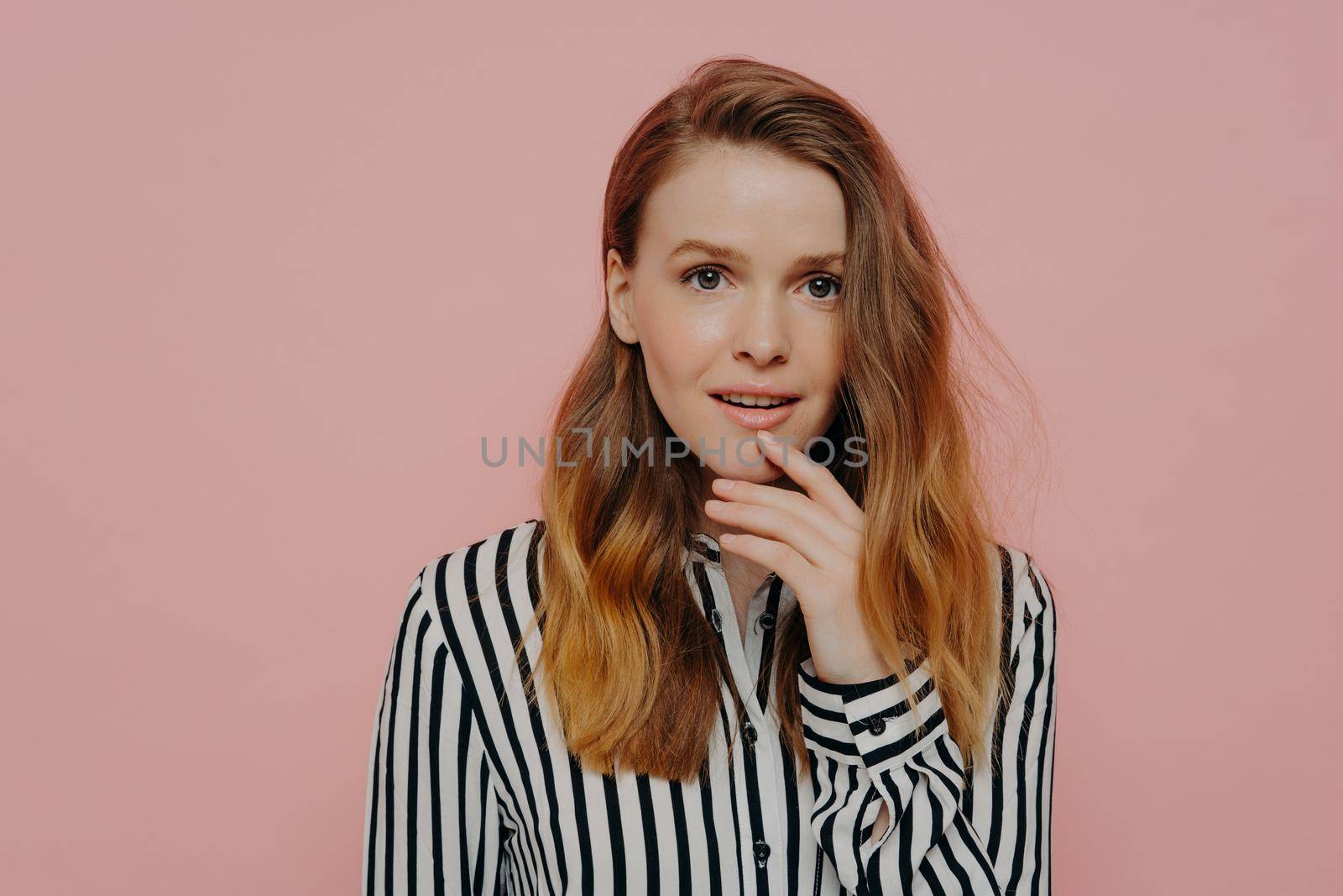 Beautiful young female student dressed in striped shirt touching chin and can not believe her eyes, looking at camera with amazed face expression, posing on pink background in studio