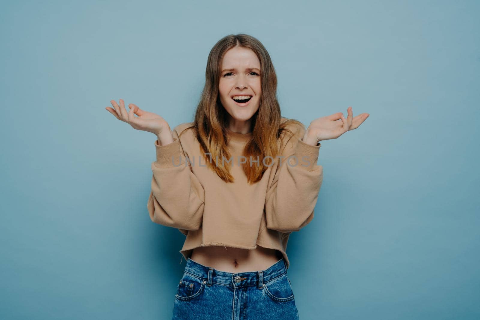 Studio shot of attractive amazed teenage girl in stylish casual outfit can not believe her eyes, shrugging shoulders with surprised face expression, looking at camera, isolated on blue background
