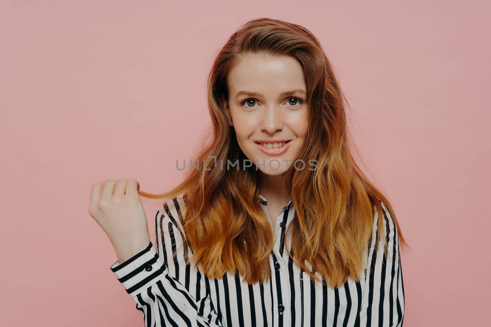 Beautiful happy young female in stripy black and white blouse holding strand of hair and smiling at camera while posing over light pink studio background