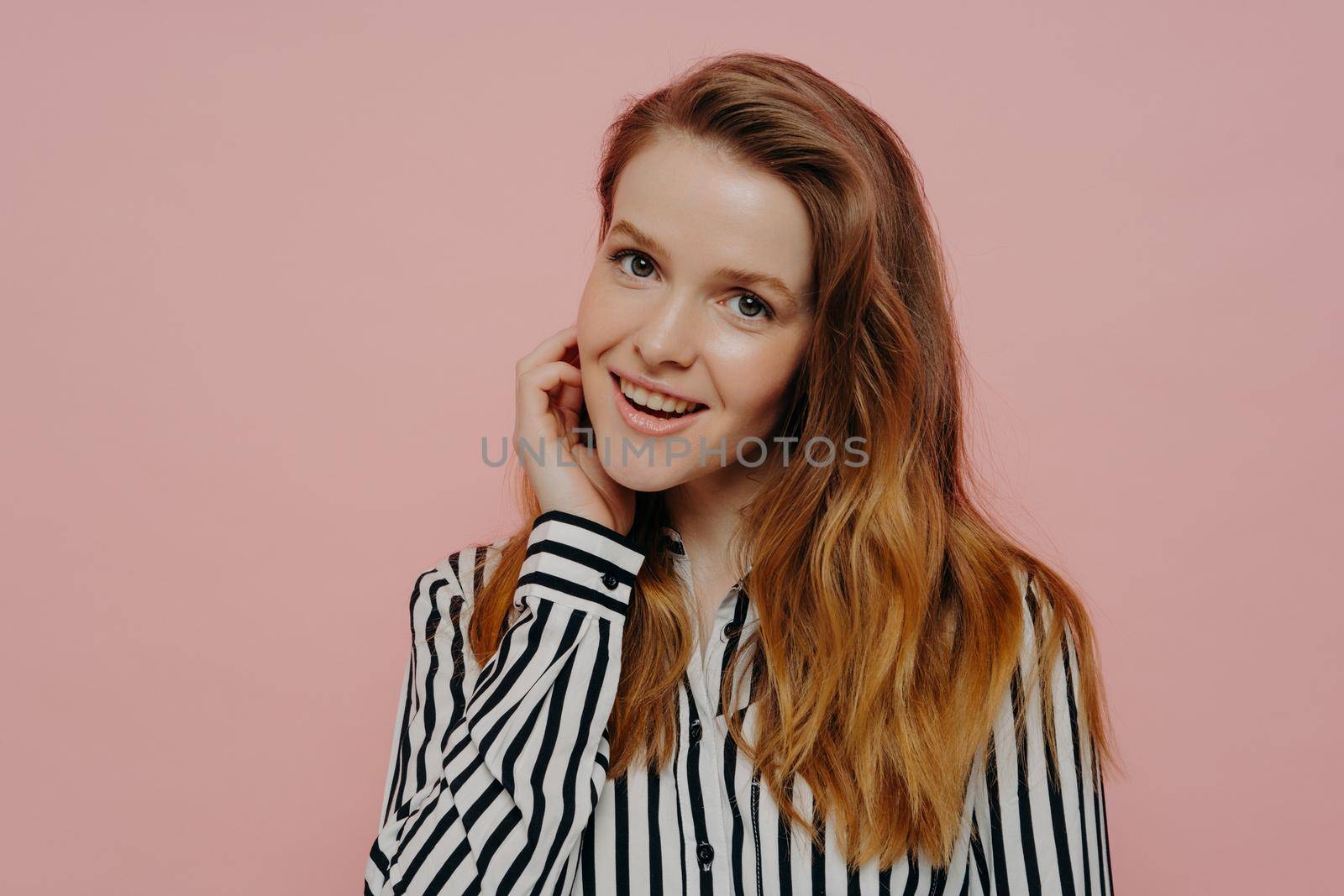 Natural beauty. Charming shy teenage girl wearing shirt holding hand near face and smiling flirty at camera while standing isolated on pink background, beautiful female posing in studio
