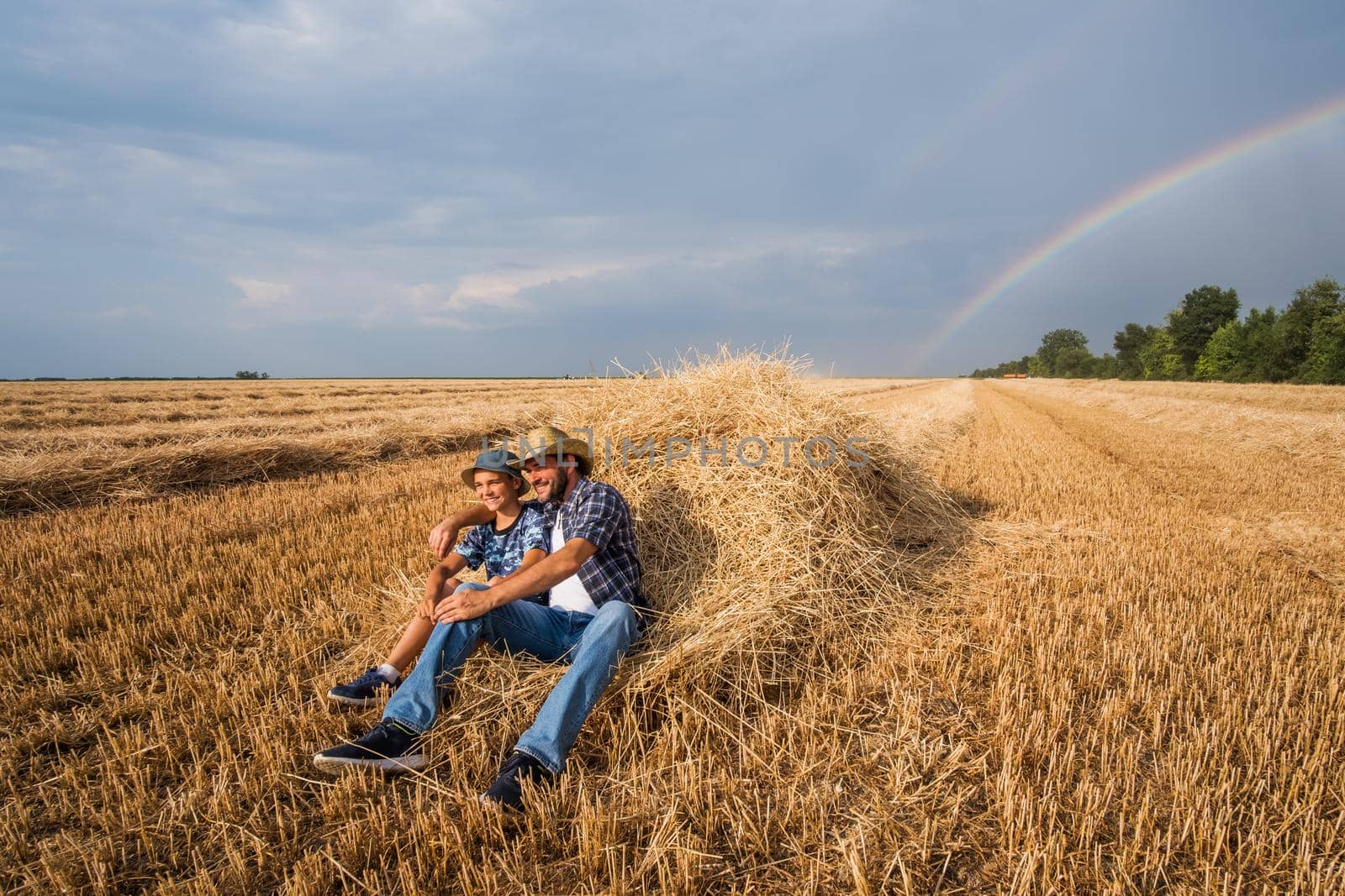 Father and son are resting after successful harvest. Rainbow in the sky behind them.