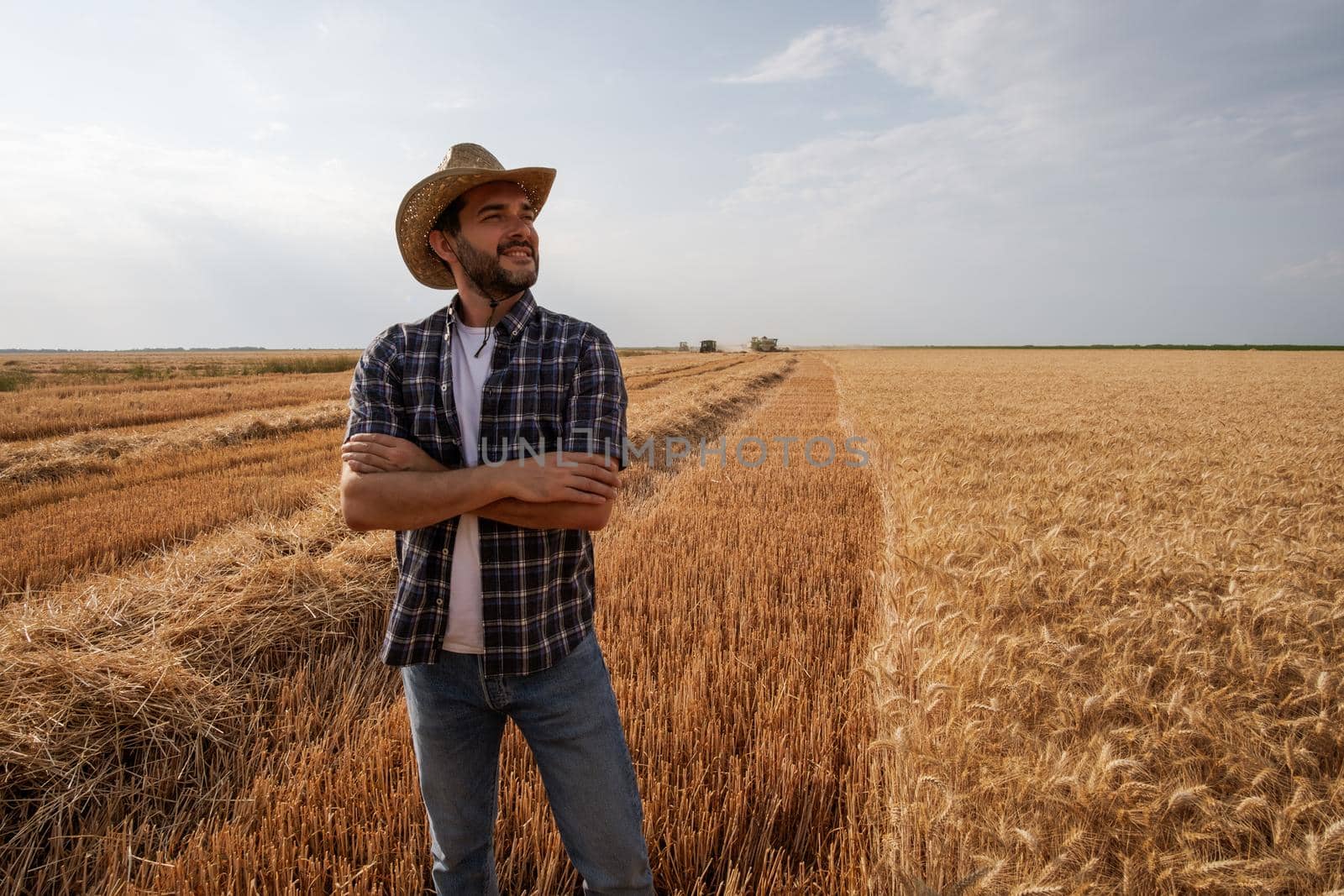 Farmer is standing in front of his wheat field while harvesting is taking place.