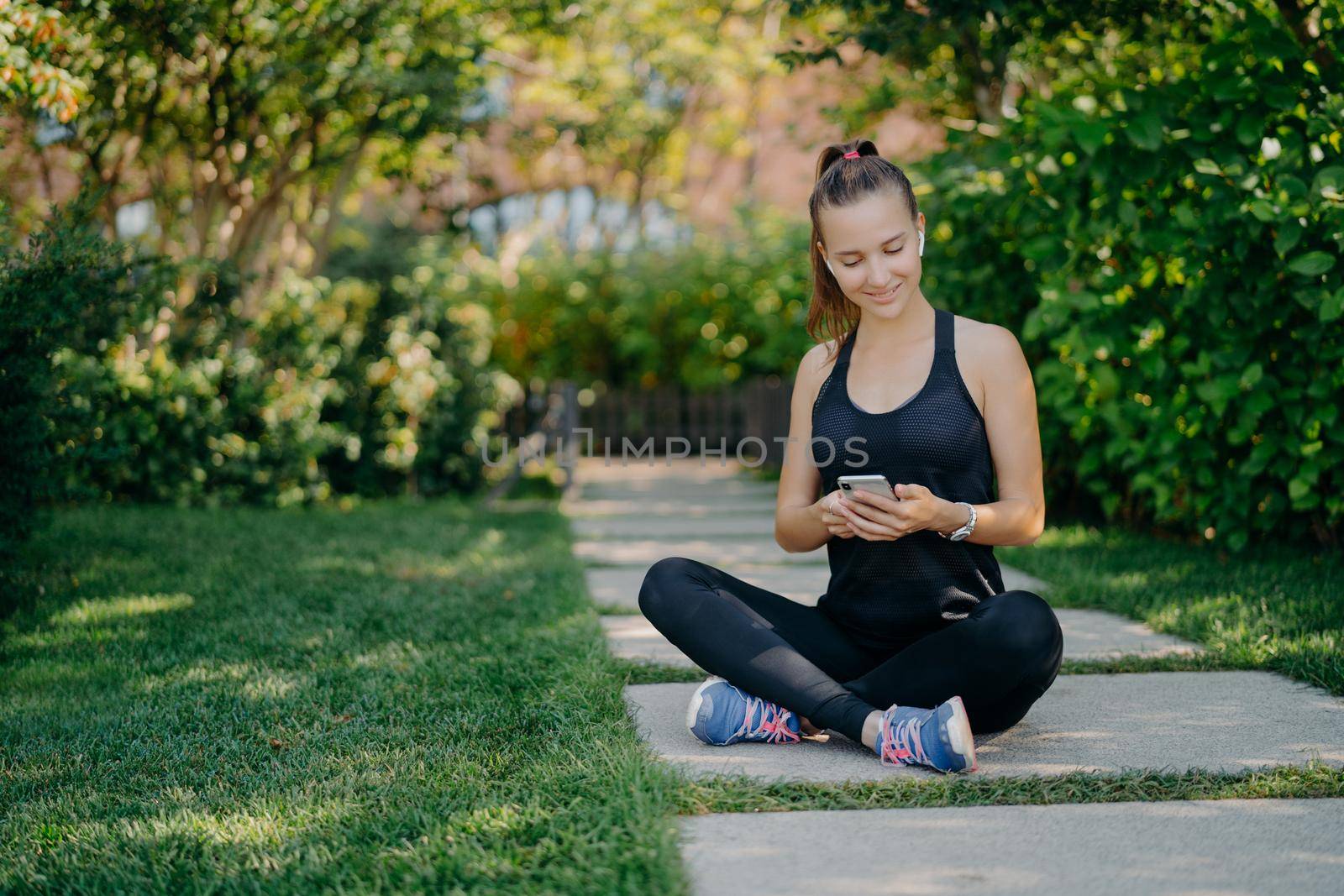 Sporty woman has break for refreshment checks messages and notification shares content about fitness outdoor dressed in active wear leads healthy lifestyle relaxes after active cardio training
