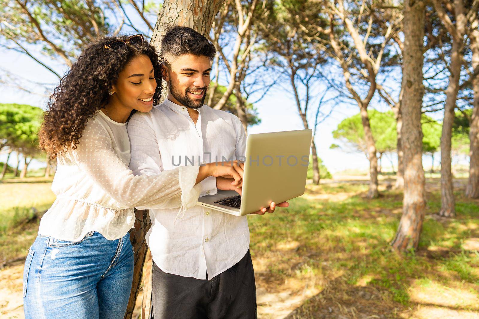 Young multiracial couple having fun with technology in vacation standing in a pine forest in ocean resort near the sea using laptop. Afro-American black girl pointing to computer screen in nature