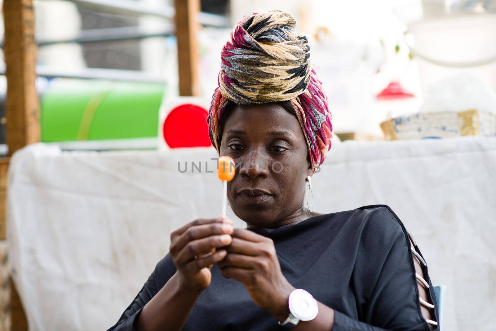 Woman with head scarf sitting behind her sales stand is holding a licked lollipop in her hand.