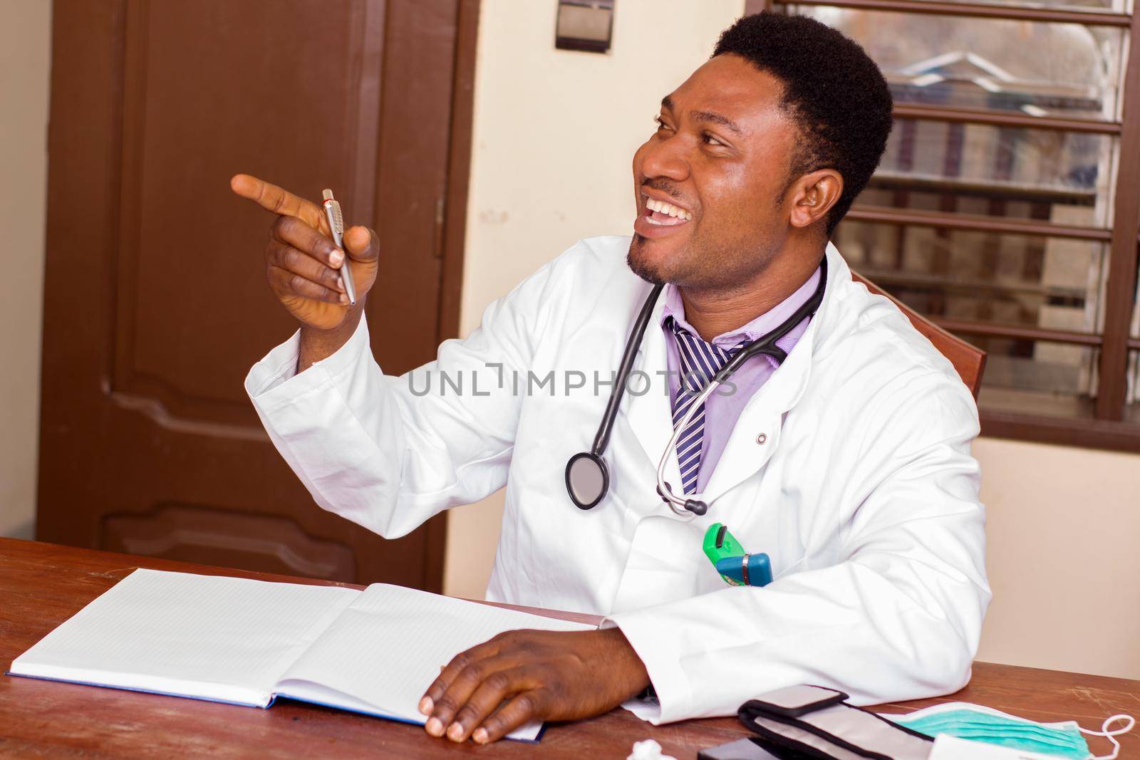 Young smiling doctor sitting at desk, talking and indexing someone in a corner of his office