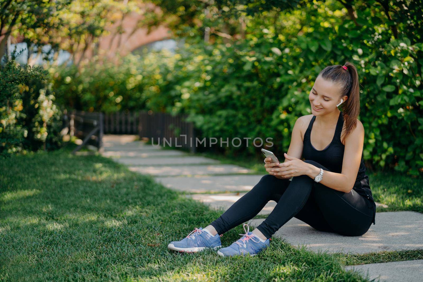 Pleased athletic woman in sportsclothes smiles cheerfully surfs mobile phone while listens music via wireless earphones takes break after sunny morning workout uses app for activity tracking
