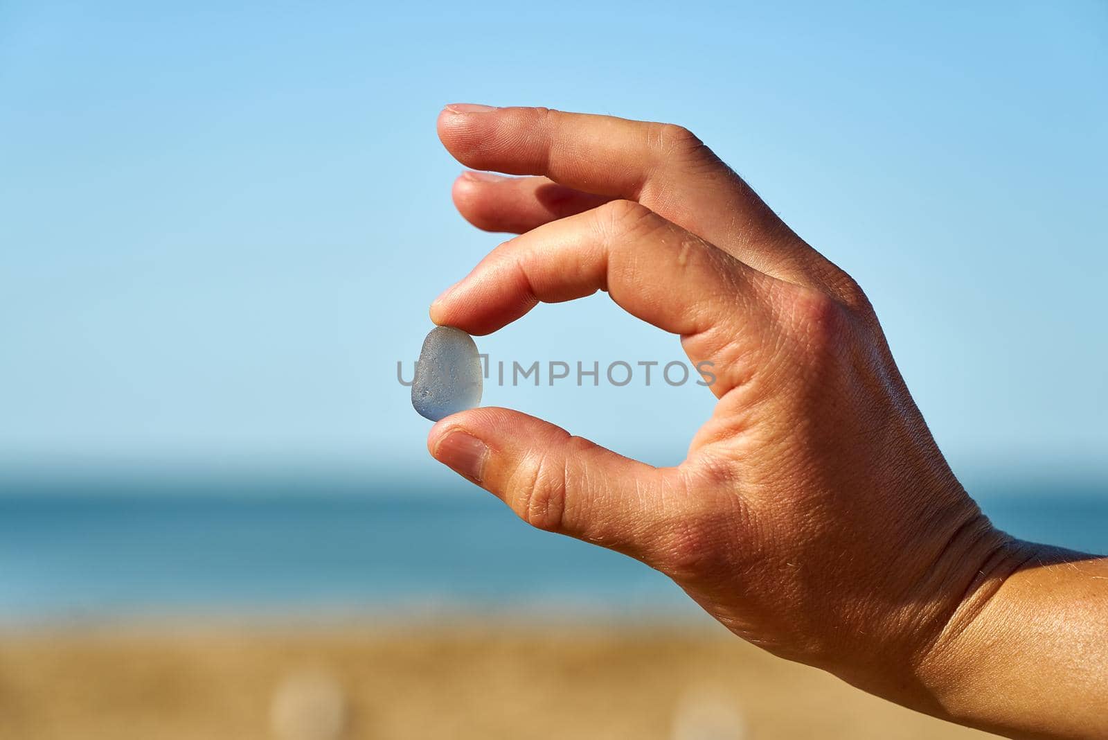 A blue piece of sea glass is held up with the sand, sea and sky in the background.