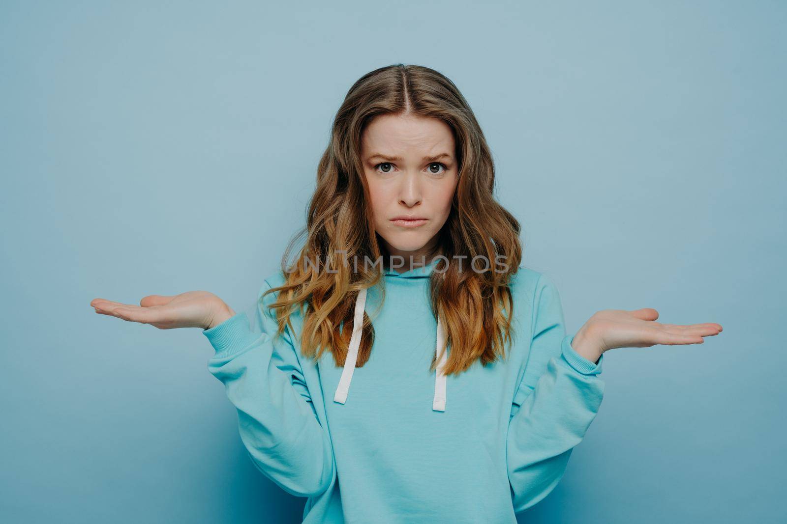Confused young female in casual hoodie cannot make decision, spreading arms with facial expression of doubt and hesitation, sad teenage girl expressing uncertainty while posing against blue background